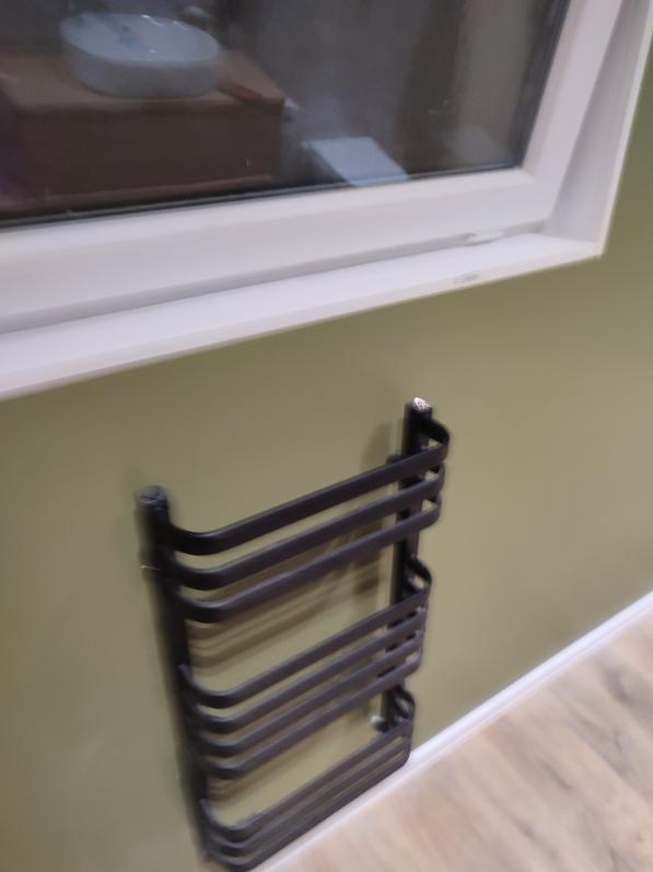 Czarny, metalowy grzejnik drabinkowy zamontowany na oliwkowozielonej ścianie łazienki, widoczny fragment okna z białymi ramami i drewnopodobna podłoga.
