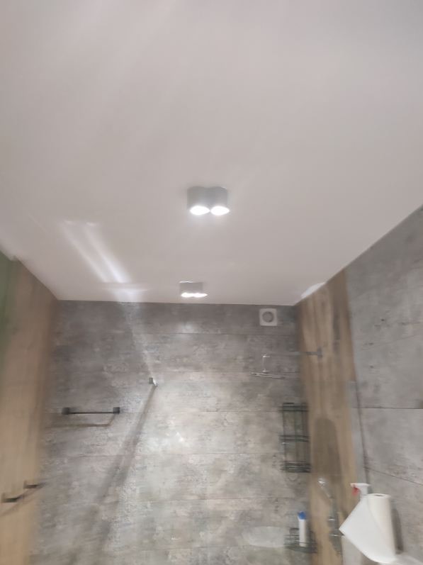 Szara łazienka z oświetleniem sufitowym, widoczne płytki imitujące beton, minimalistyczne półki i wieszaki na ręczniki.