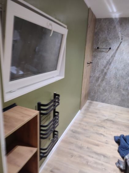 Uchylone okno PCV w łazience z zieloną ścianą, obok drewniany regał i metalowy wieszak na ręczniki; podłoga wyłożona panelami imitującymi drewno, w tle imitacja kamienia na ścianie.