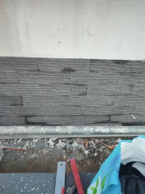 Fragment ściany z szarej, wąskiej okładziny kamiennej w trakcie montażu, widoczne niedociągnięcia i narzędzia glazurnicze u dołu.