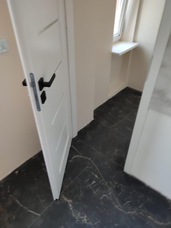 Ciemna, marmurkowa glazura podłogowa w korytarzu, widoczny fragment białych drzwi z czarną klamką i parapet okienny.