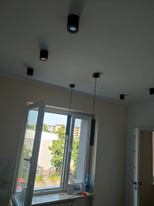 Wnętrze pokoju z otwartym oknem i zamontowanym oświetleniem sufitowym: czarne lampy punktowe i wiszące cylindryczne oprawy oświetleniowe.