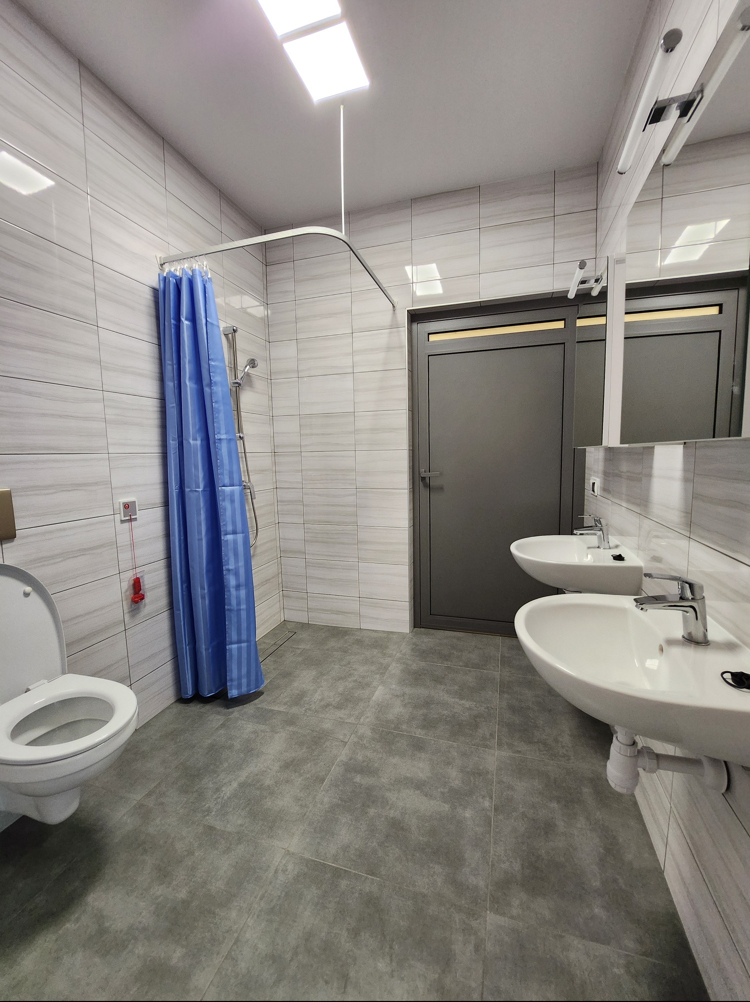 Nowoczesna łazienka z szarymi płytkami, dwoma umywalkami, toaletą i zasłoną prysznicową w kolorze niebieskim, wyposażona w system alarmowy na ścianie.