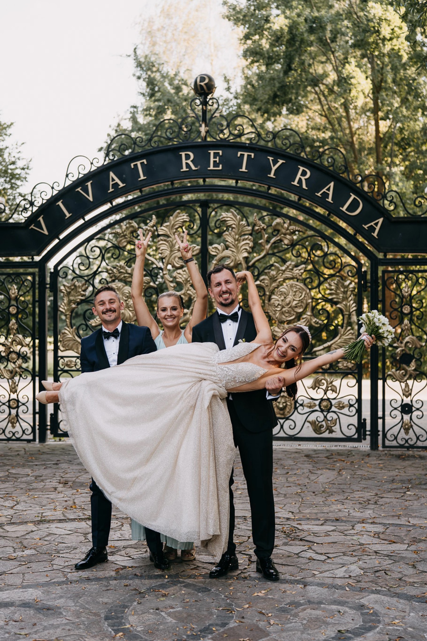 Uśmiechnięta panna młoda w sukni ślubnej niesiona przez dwóch mężczyzn w smokingach przed ozdobną bramą z napisem 'Vivat Retyrada', w tle kobieta z podniesionymi rękami.