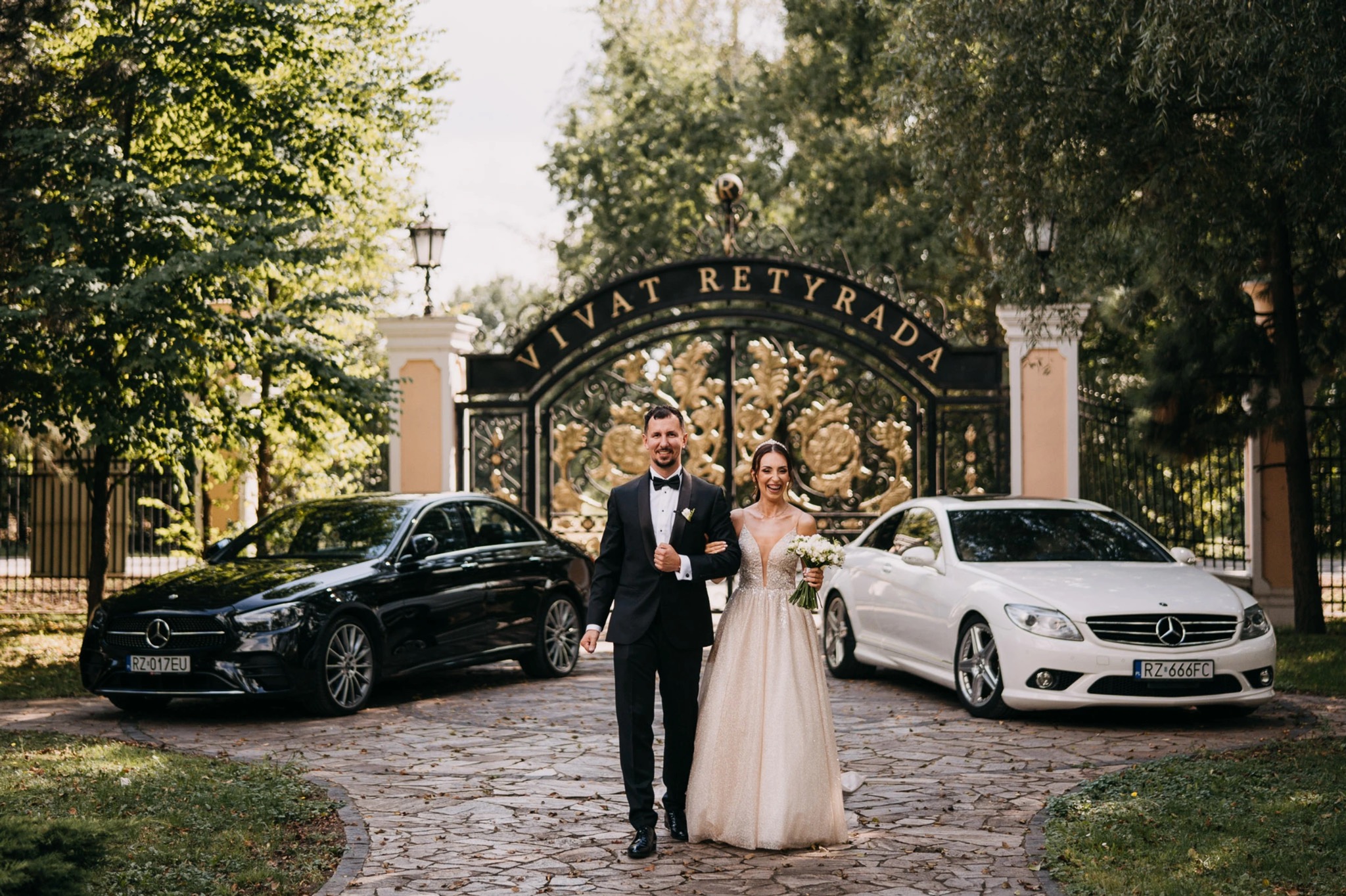 Uśmiechnięta para młoda idzie aleją w kierunku kamery, obok stoją dwa luksusowe samochody, brama z napisem 'Vivat Retyrada' w tle.