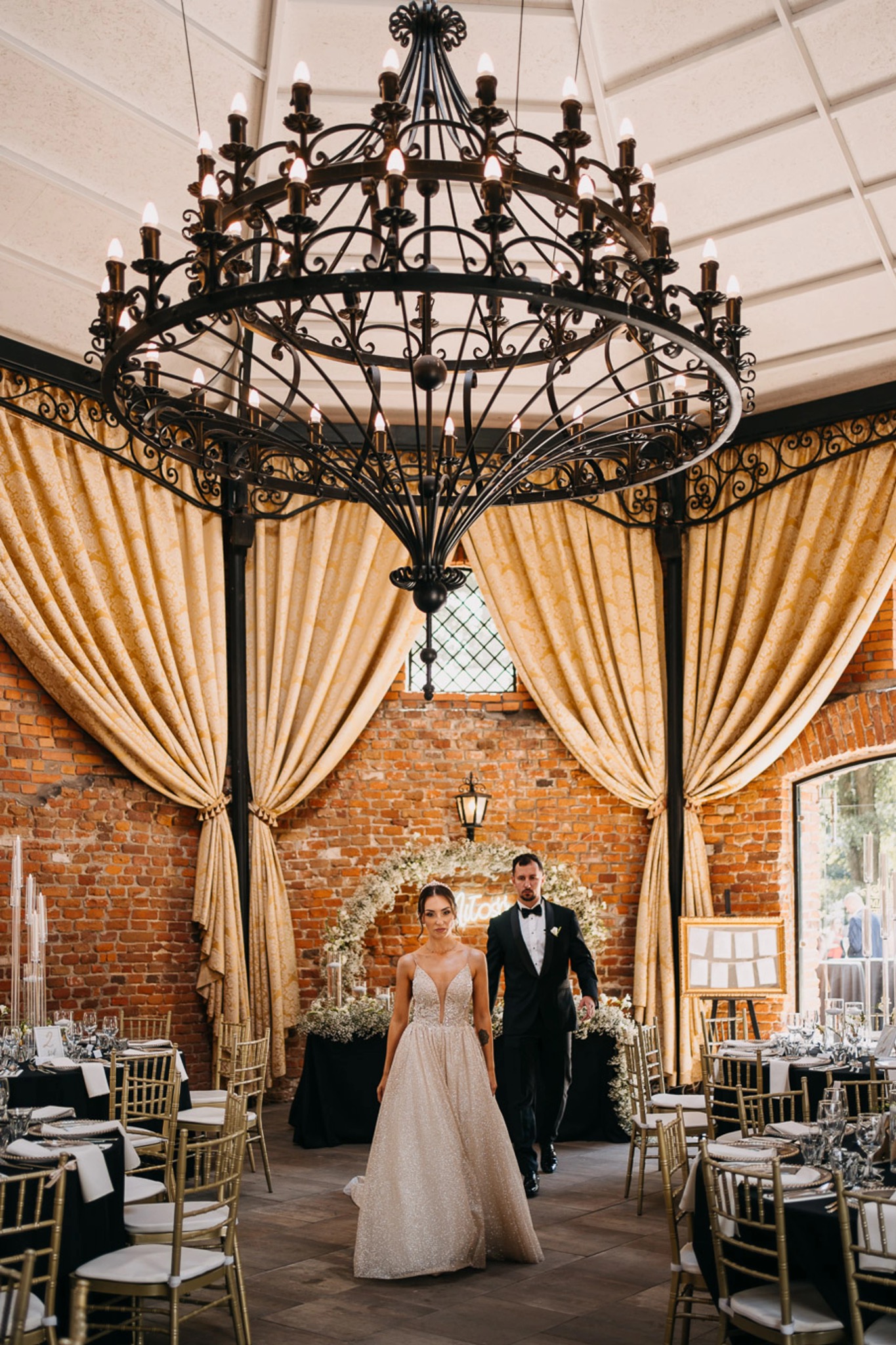 Para młoda w eleganckim wnętrzu sali weselnej z ceglanymi ścianami, dekoracjami kwiatowymi i żyrandolem w stylu retro.