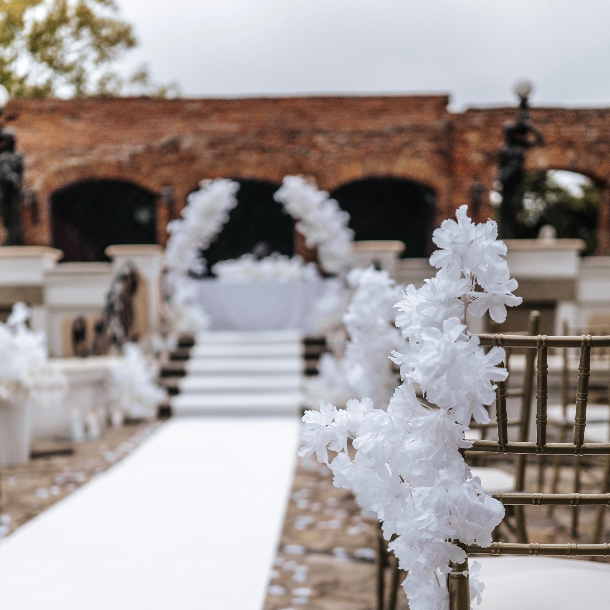 Dekoracja krzesła białymi kwiatami na ślubnej ceremonii plenerowej z białym dywanem i ceglanym budynkiem w tle.