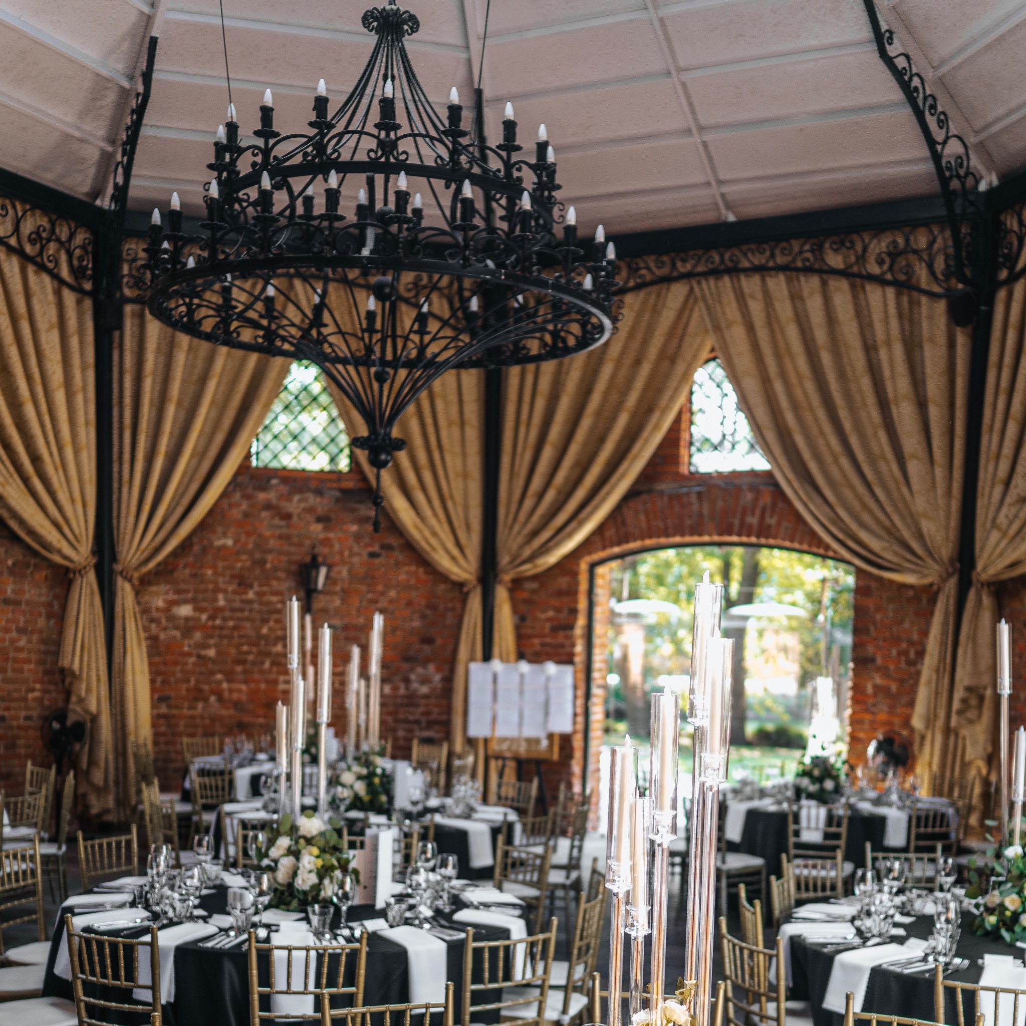 Elegancka sala bankietowa z dekoracjami w czerni i bieli: okrągłe stoły z złotymi krzesłami, szklane świeczniki i kwiatowe kompozycje, oświetlona żyrandolem z kutego żelaza.