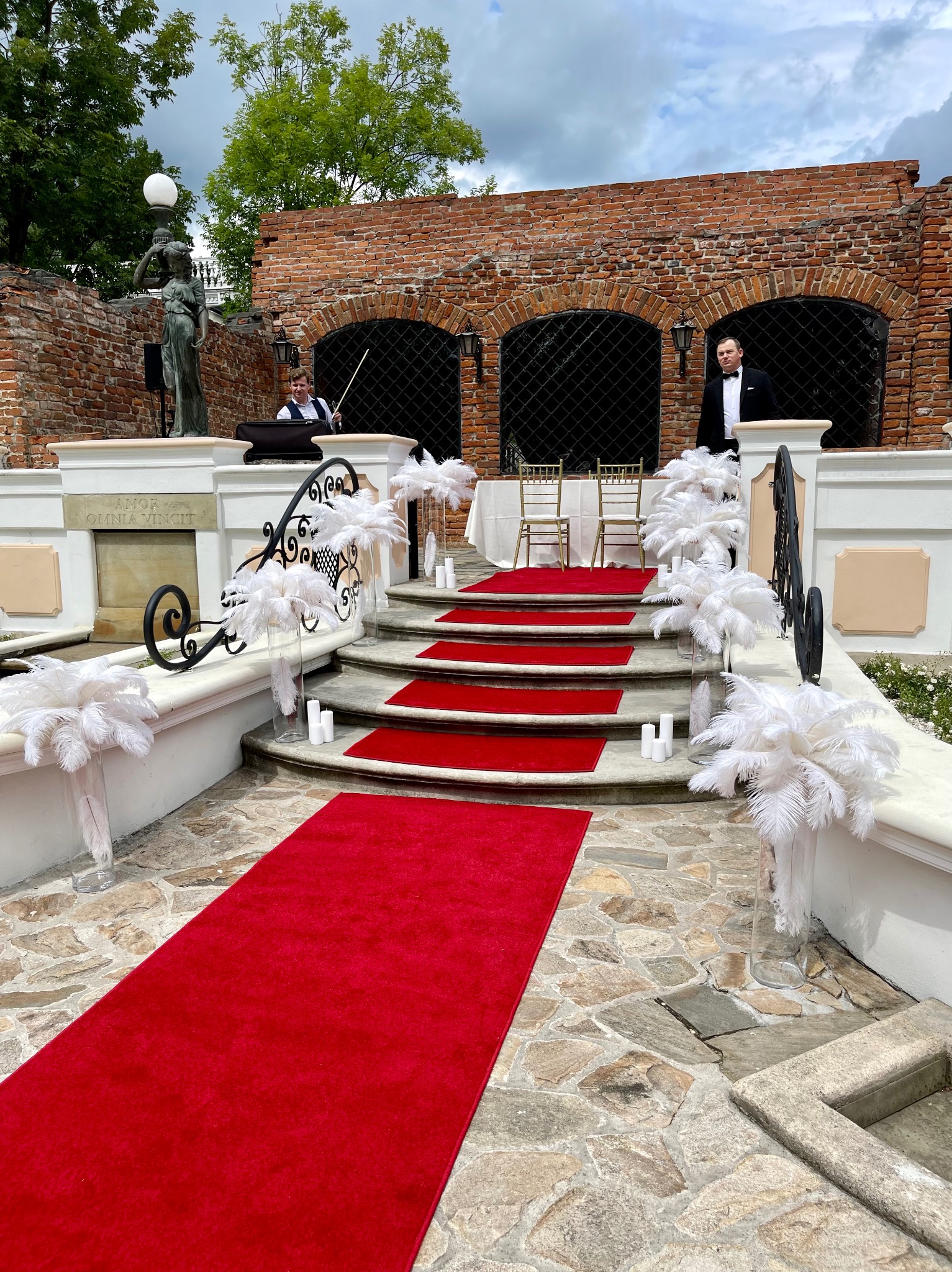 Uroczyste wejście po schodach z czerwonym dywanem, udekorowane białymi piórami w wazonach i świecami, z muzyką na żywo w tle.