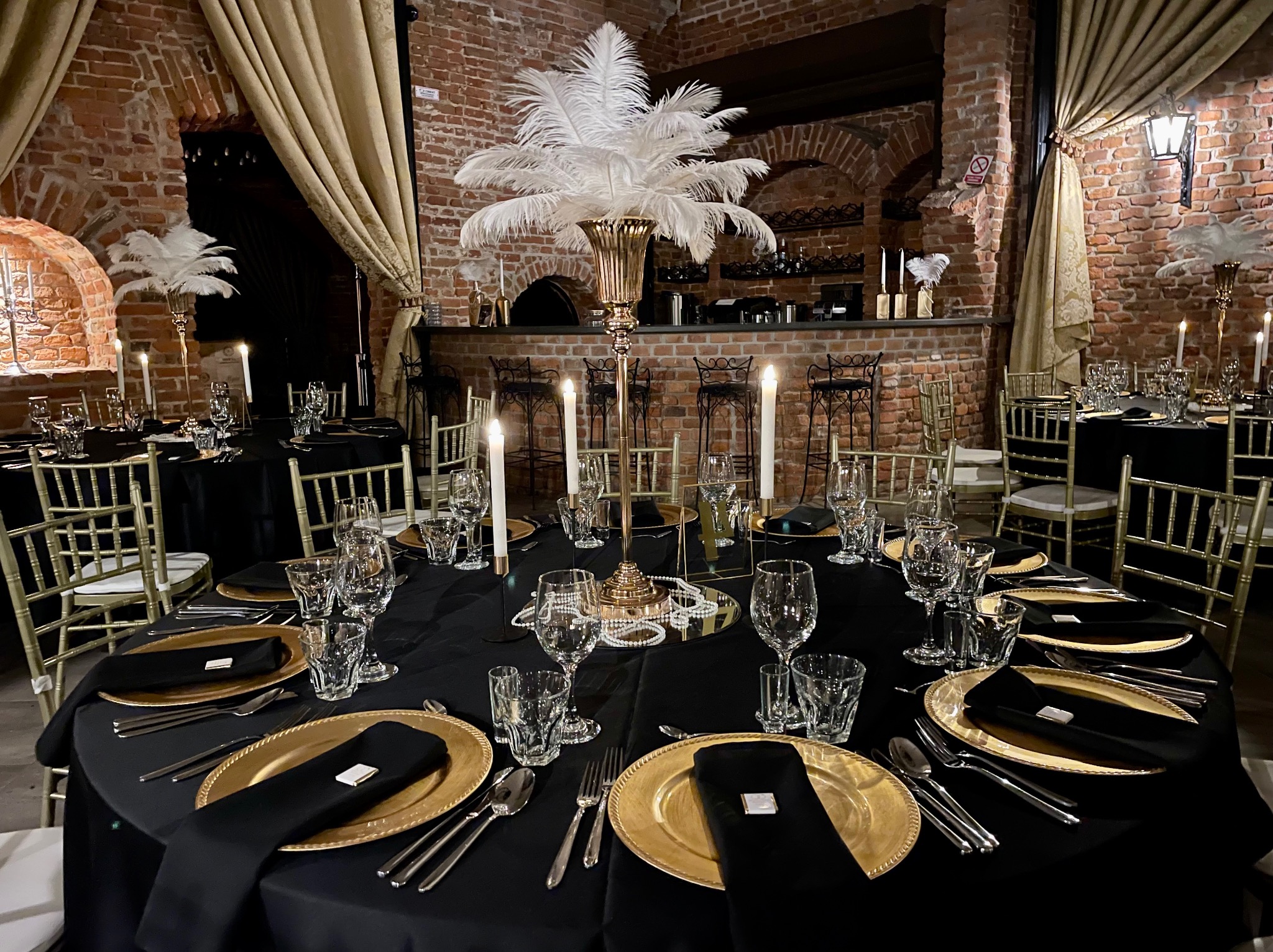 Elegancko nakryty stół z czarnym obrusem, złotymi talerzami i białymi piórami w wysokich wazonach, w rustykalnym wnętrzu z ceglanymi ścianami i barem w tle.