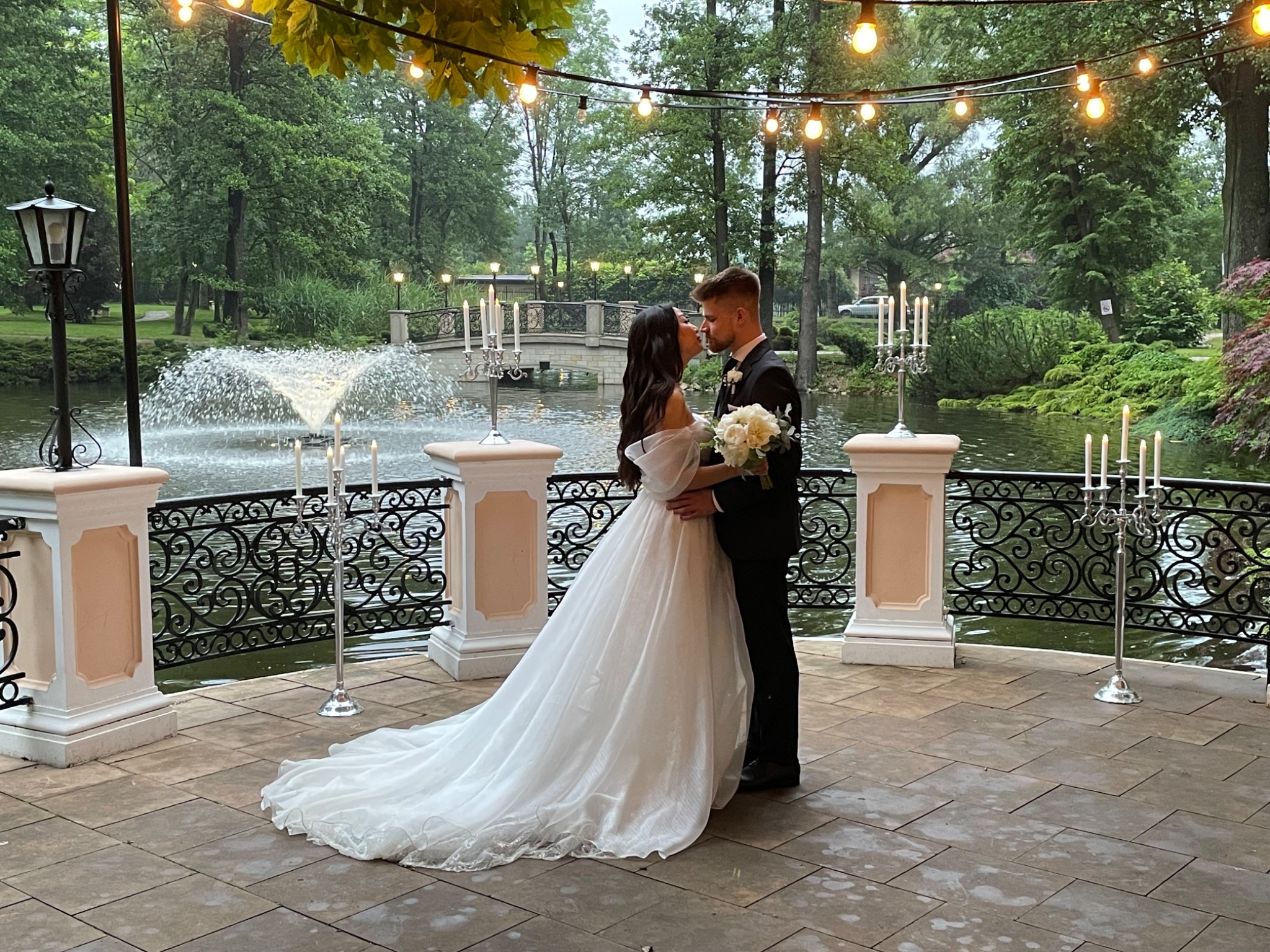 Para młoda w objęciach na tle fontanny, dekoracji ze świec i girland z żarówek w plenerowej scenerii.