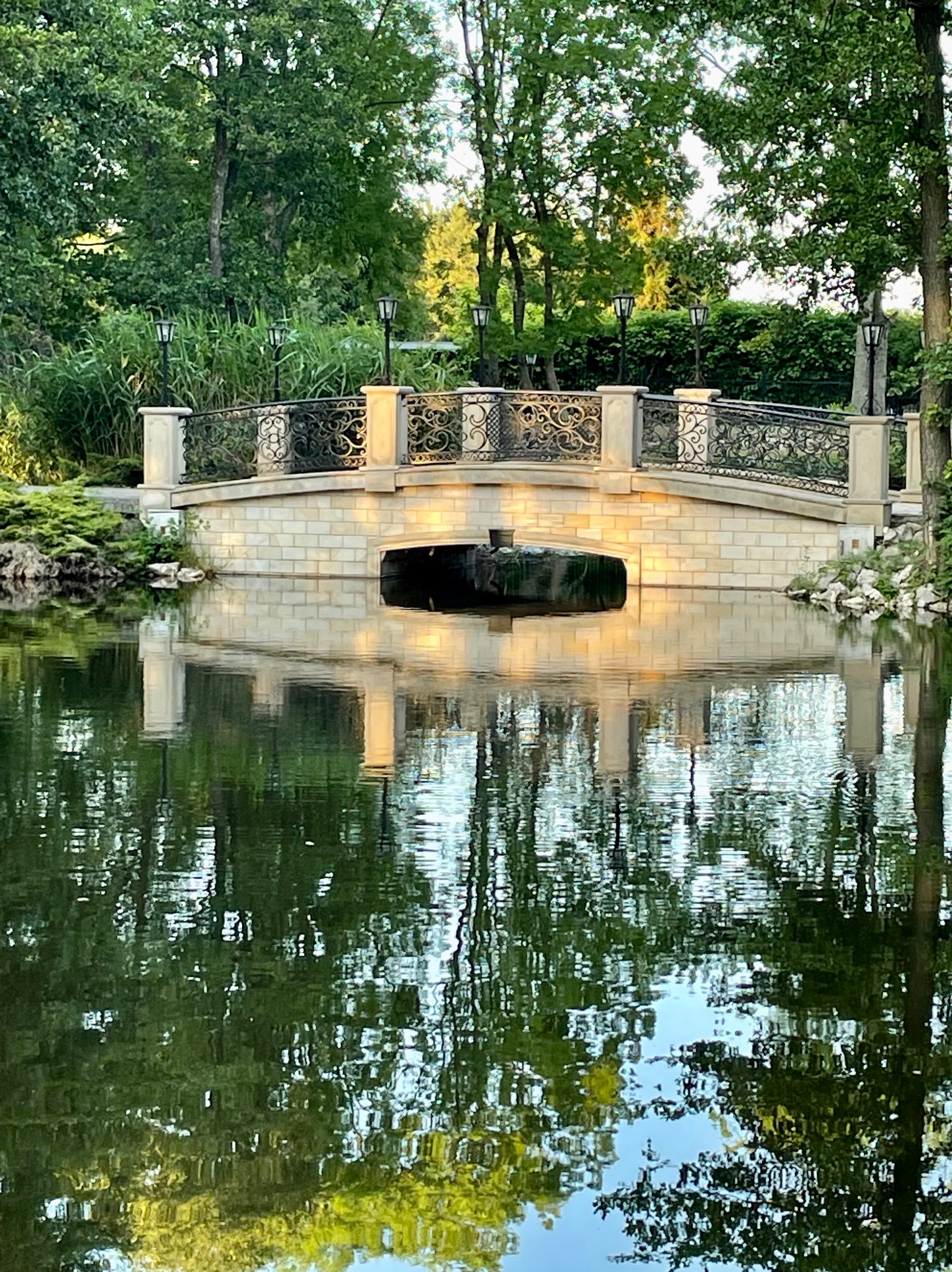 Kamienny mostek z ozdobną, metalową balustradą nad stawem, oświetlony lampionami, z wyraźnym odbiciem w wodzie.