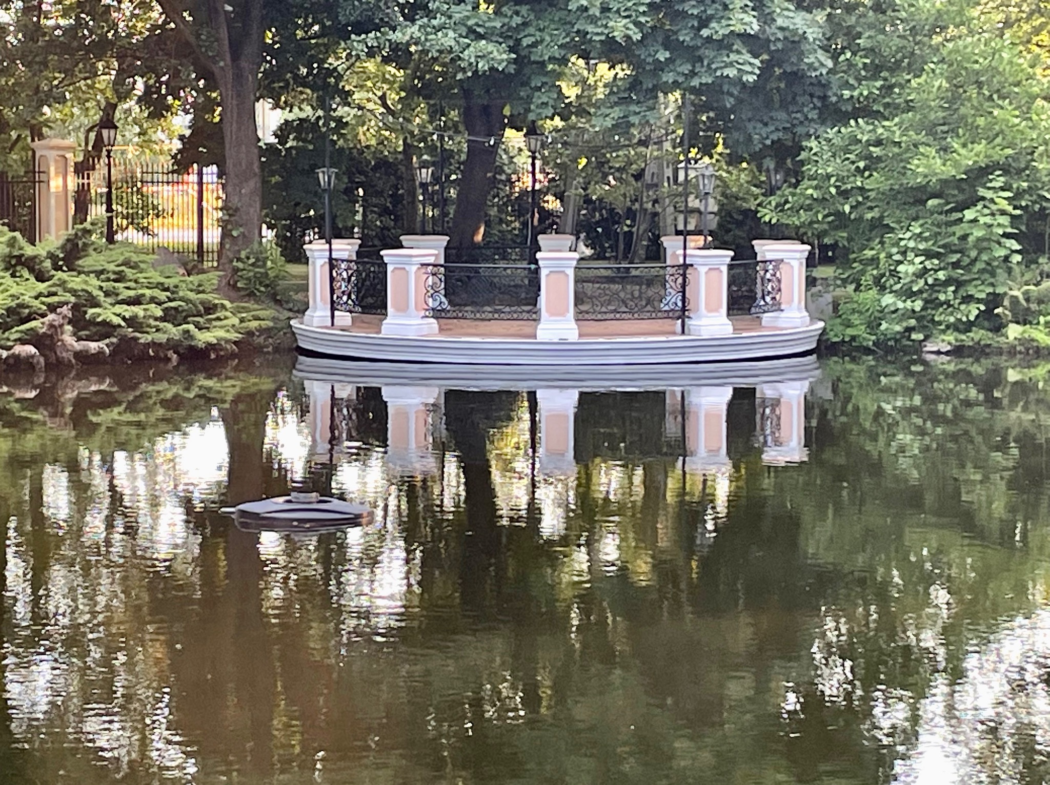 Okrągła platforma z kolumnami i ozdobnymi balustradami na stawie, otoczona zielenią, z widocznym odbiciem w wodzie.