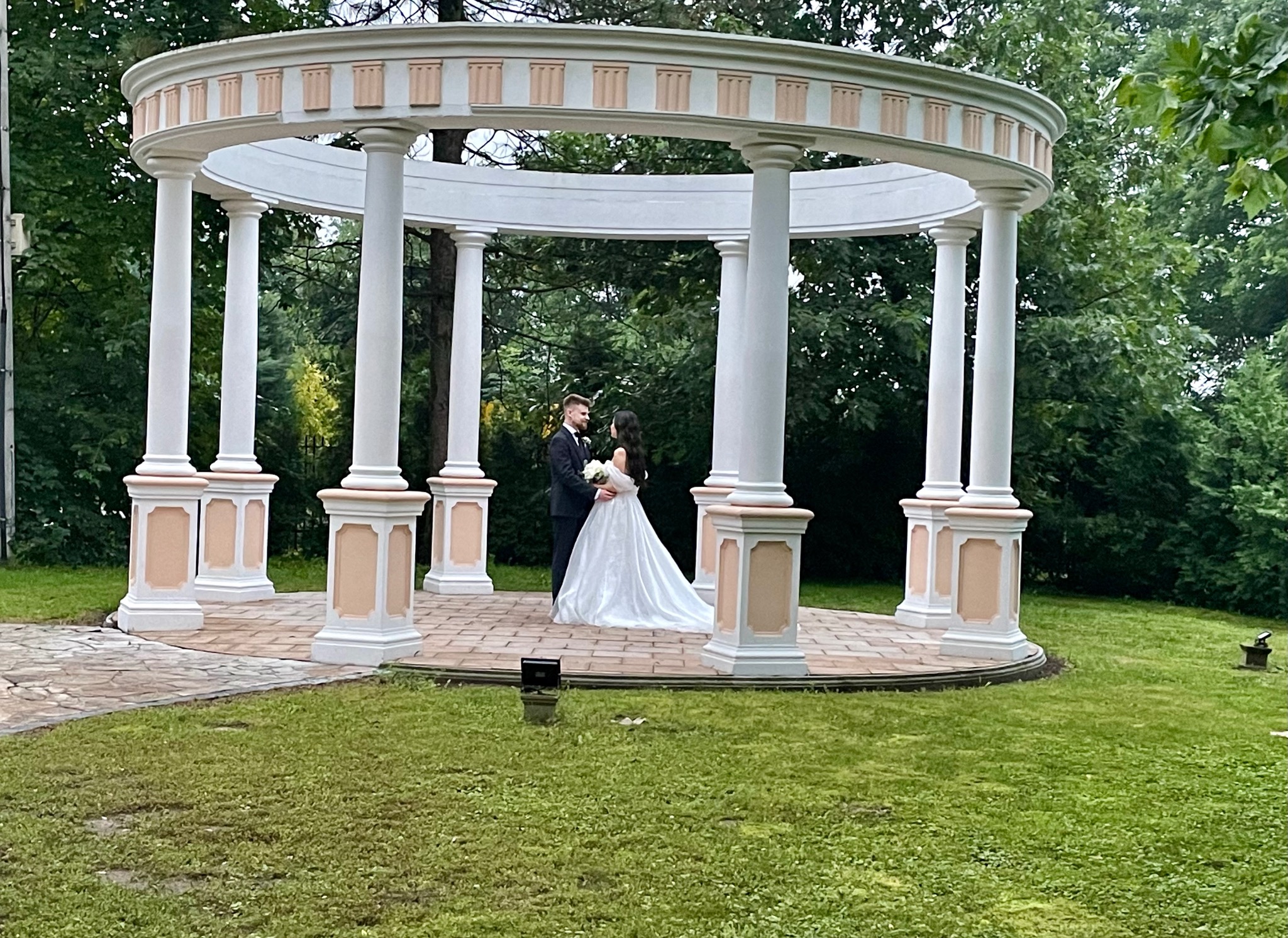Para młoda w białej sukni i czarnym garniturze stoi w okrągłej, kolumnowej altanie z kremowymi akcentami, otoczonej zielenią drzew i trawnikiem.