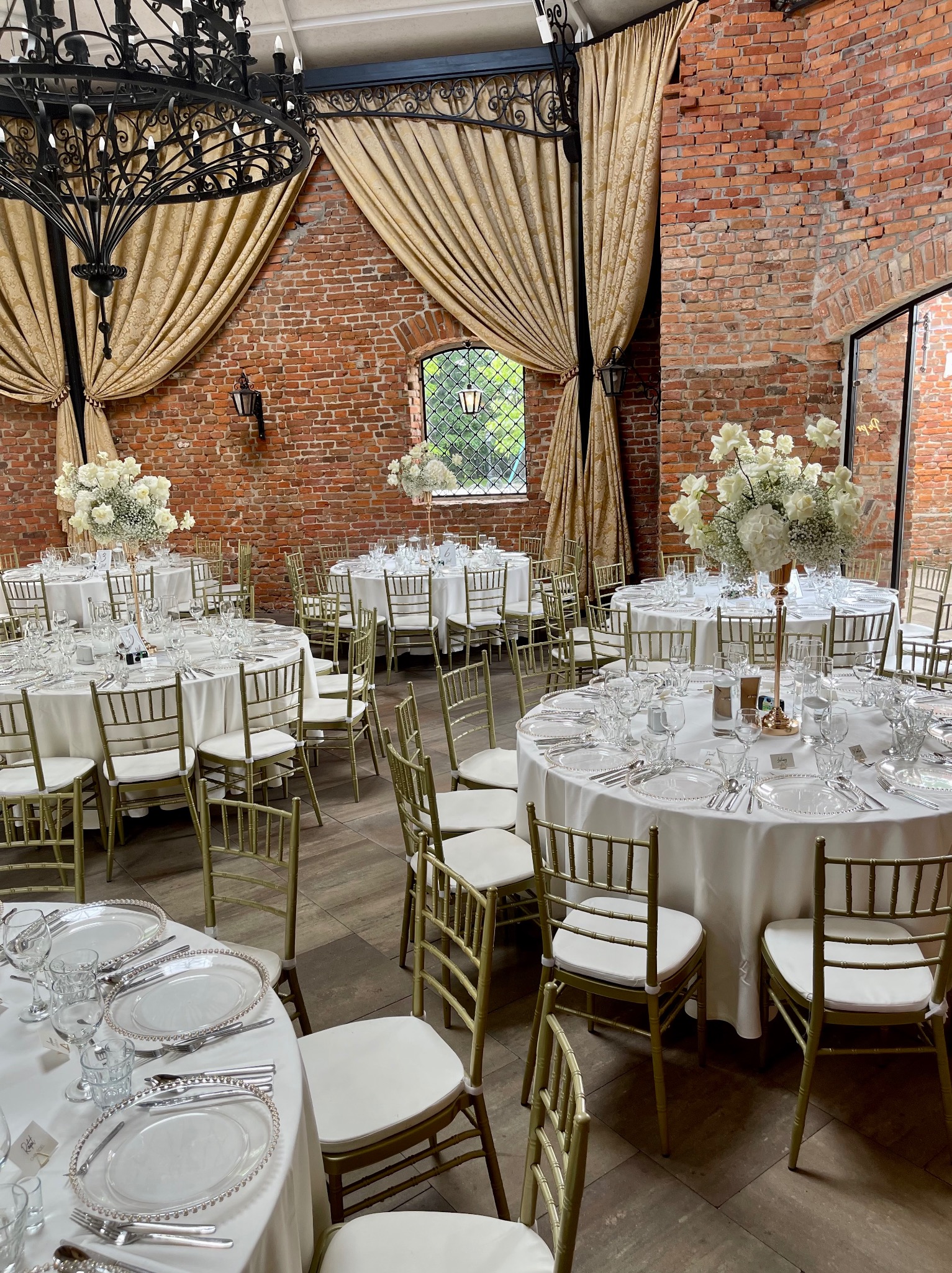 Sala weselna z okrągłymi stołami nakrytymi białymi obrusami, złotymi krzesłami, elegancką zastawą i wysokimi kompozycjami z białych kwiatów, w tle ceglane ściany i złote zasłony.