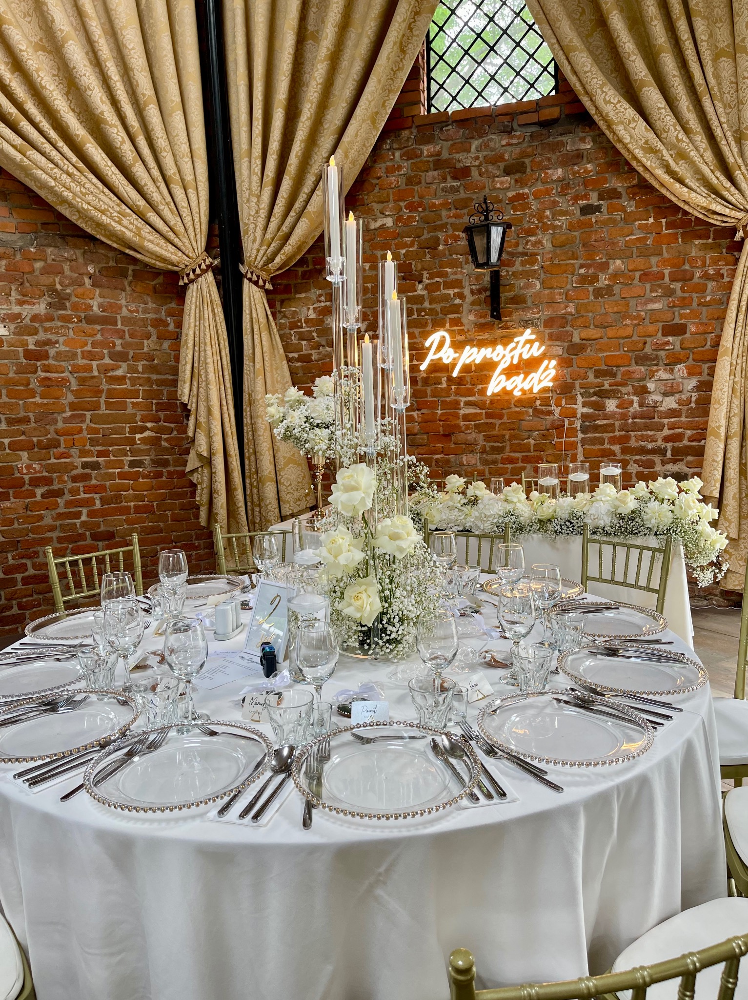Uroczysty stół weselny nakryty białym obrusem, z dekoracjami z białych róż i gipsówki, szklanymi świecznikami z długimi świecami, ozdobnymi talerzami i sztućcami, na tle ceglanej ściany z neonowym...