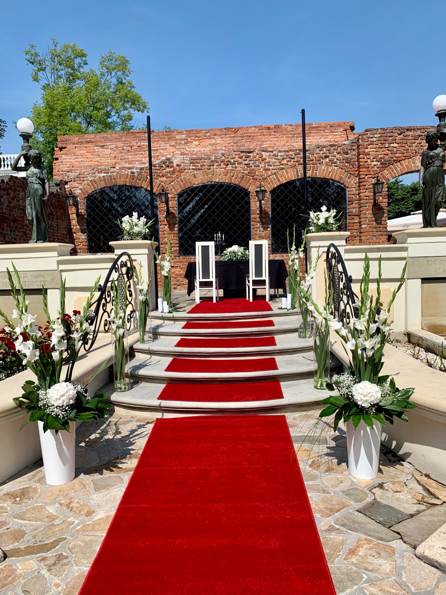 Uroczysta aranżacja ślubna na zewnątrz: czerwony dywan prowadzi po schodach do stołu z białymi krzesłami, otoczonego kwiatowymi dekoracjami w białych wazonach, na tle ceglanej ściany z łukowymi...