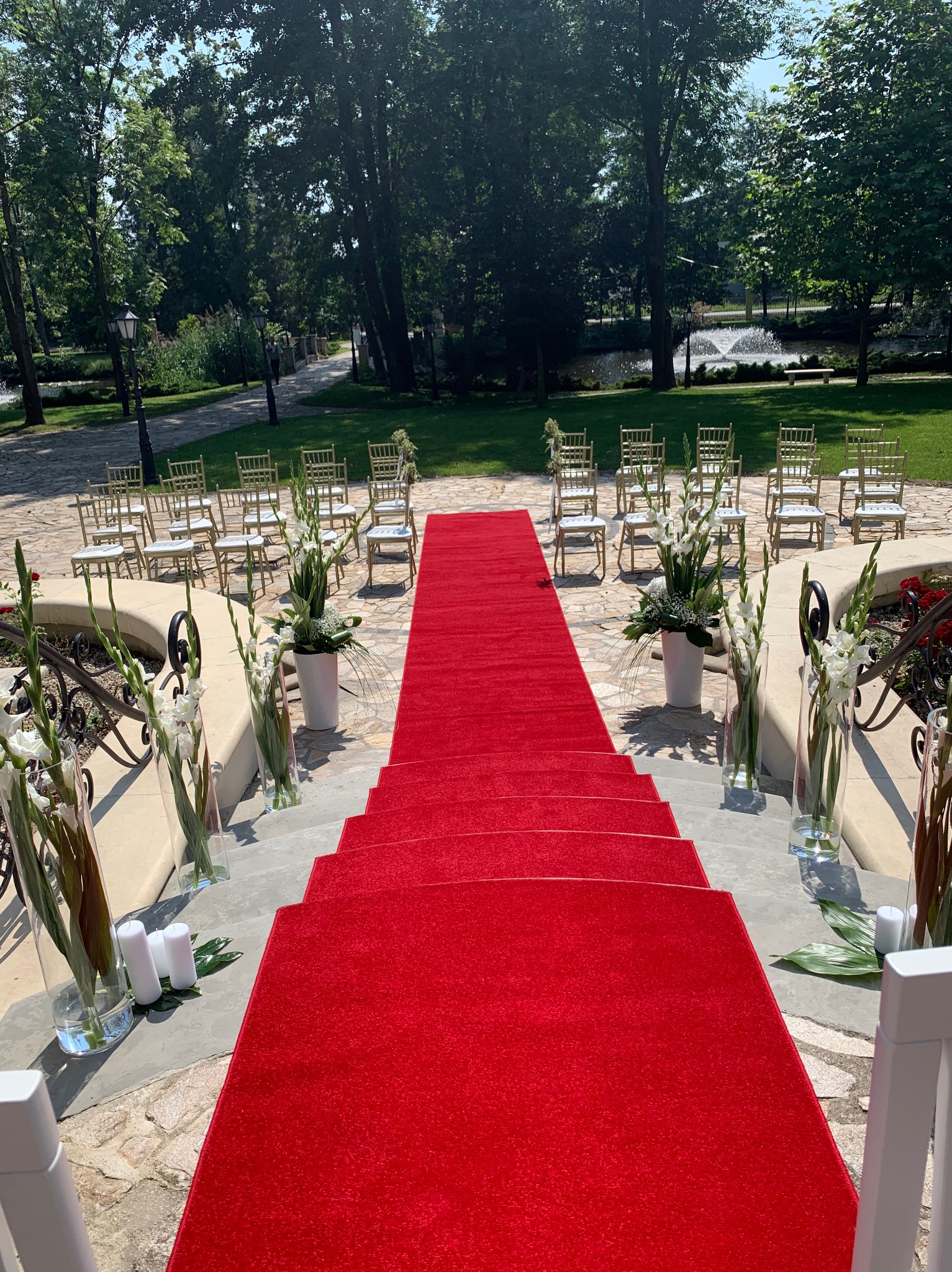 Czerwony dywan na schodach prowadzący do rzędu krzeseł na zewnątrz, dekoracje kwiatowe w szklanych wazonach i białe świece, tło park z fontanną.