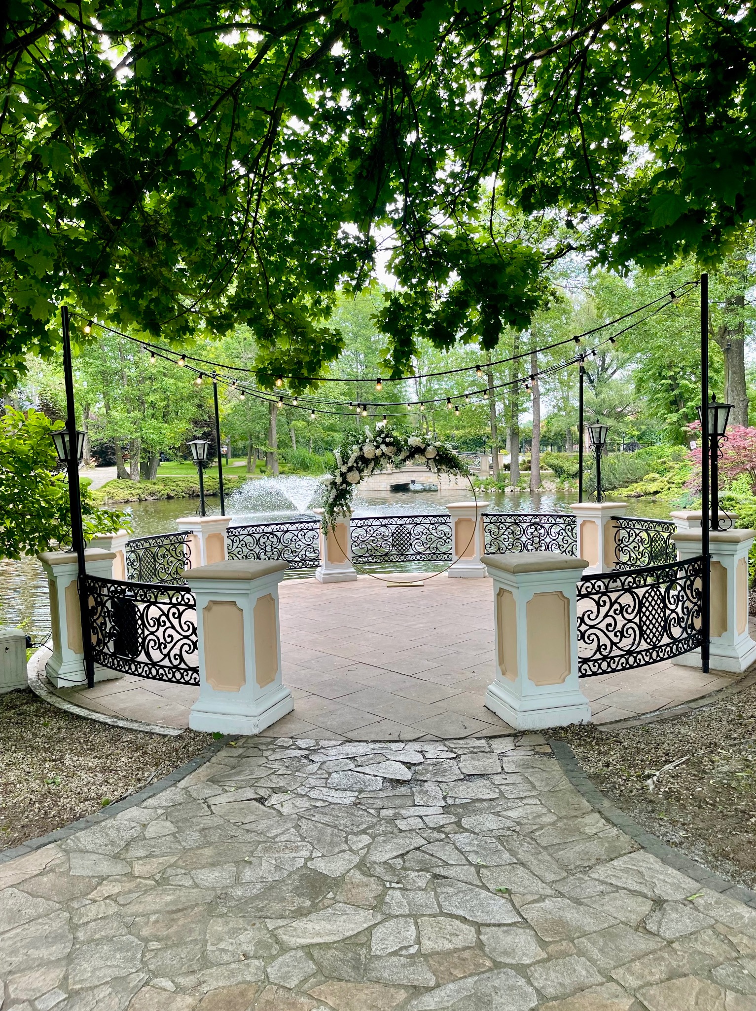 Dekoracja ślubna w plenerze: okrągła brama z kwiatami nad stawem, oświetlona girlandami żarówek, widok z kamiennej ścieżki.