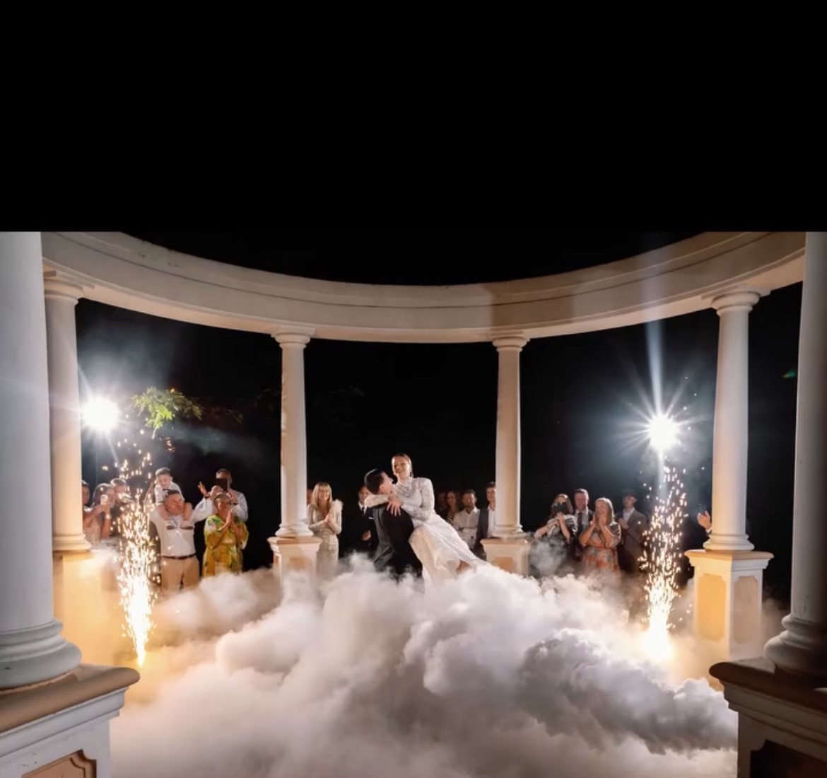 Para młoda w objęciach, unoszona w dymie i blasku fontann iskier, otoczona gośćmi pod kolumnową altaną.