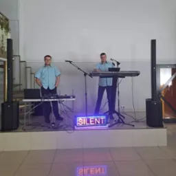 Dwaj mężczyźni w niebieskich koszulach na scenie z instrumentami muzycznymi i podświetlanym napisem 'SILENT', jeden stoi z rękami w kieszeniach, drugi gra na keyboardzie.