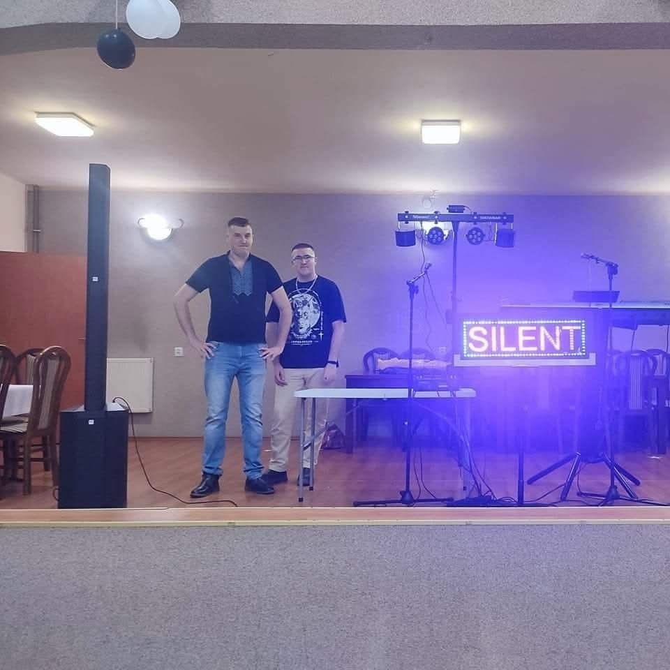 Zespół muzyczny na scenie z napisem 'SILENT' podświetlonym na niebiesko, jeden z muzyków w jeansach i koszuli z haftem, drugi w czarnej koszulce z nadrukiem i okularach.