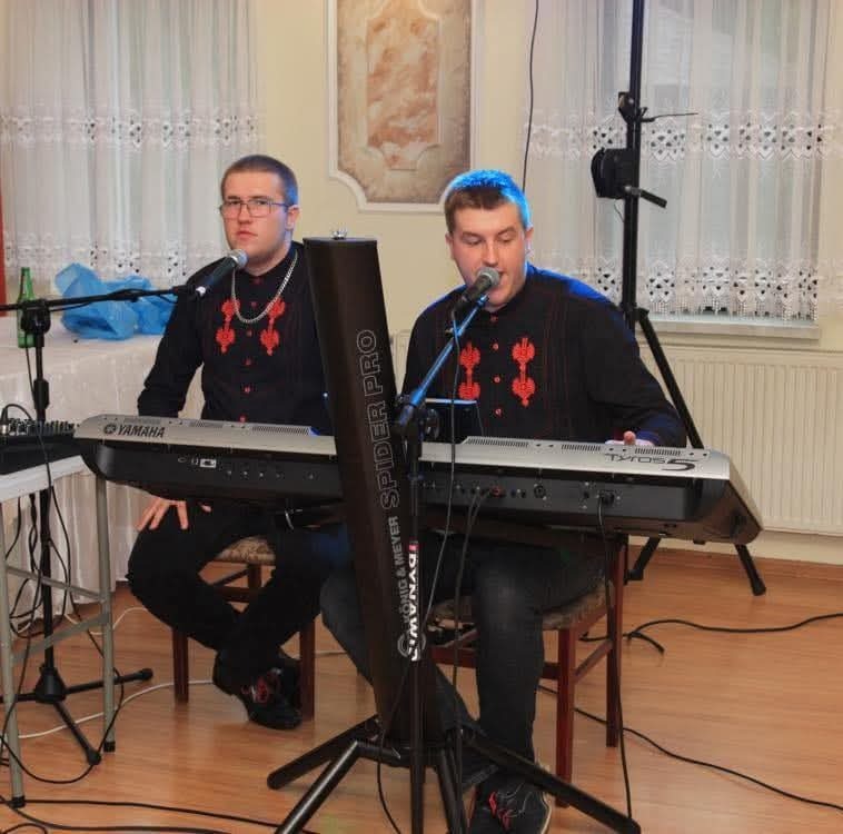 Dwóch mężczyzn w czarnych koszulach z czerwonym haftem gra na keyboardach Yamaha i Tyros 5, śpiewając do mikrofonów na statywach, w tle białe zasłony i rama obrazu.