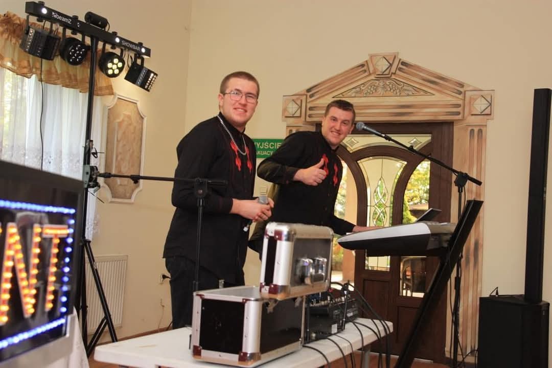 Dwóch mężczyzn w czarnych koszulach z czerwonym haftem, prezentujących się za klawiaturą i sprzętem nagłośnieniowym, jeden z nich pokazuje kciuk w górę, na tle dekoracyjnych drzwi i oświetlenia...