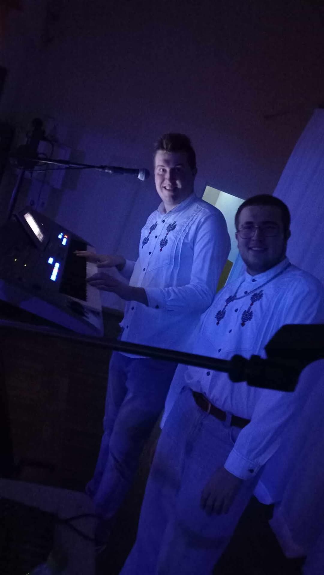 Dwóch mężczyzn w białych koszulach gra na keyboardzie podczas występu na scenie, oświetlonej niebieskim światłem.