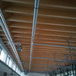 Metalowe rury wentylacyjne zamontowane pod drewnianym stropem w hali, widoczne okna i brama garażowa.