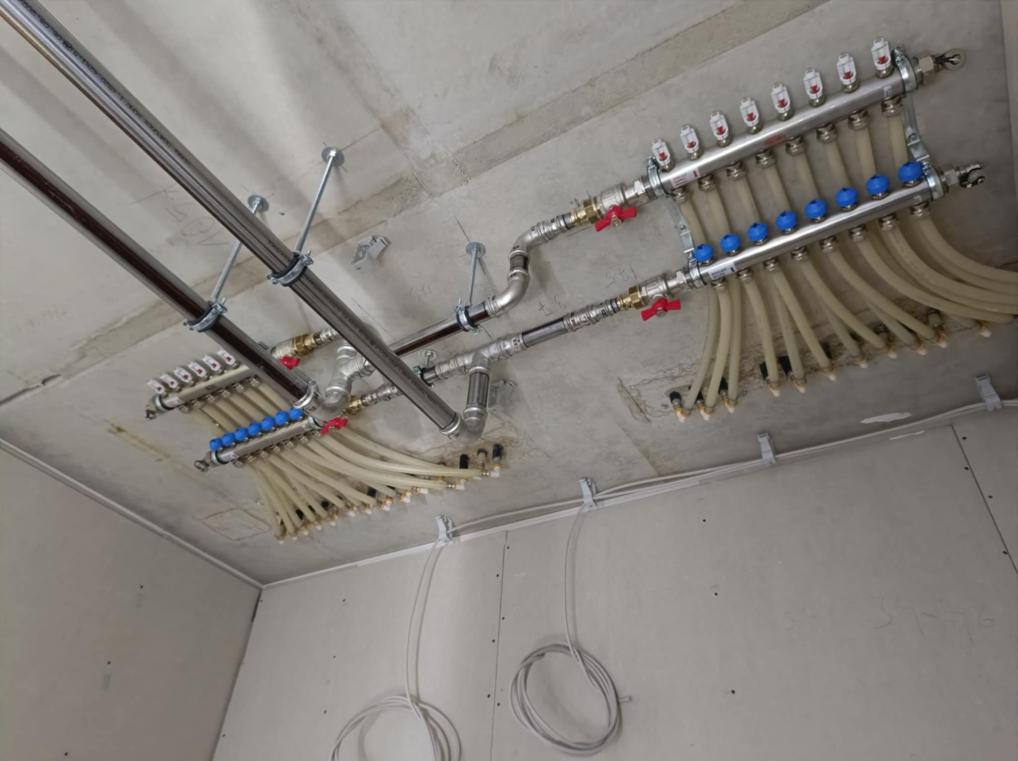Rozdzielacze ogrzewania podłogowego zamontowane pod sufitem w surowym stanie, widoczne rury i zawory, instalacja w trakcie realizacji.