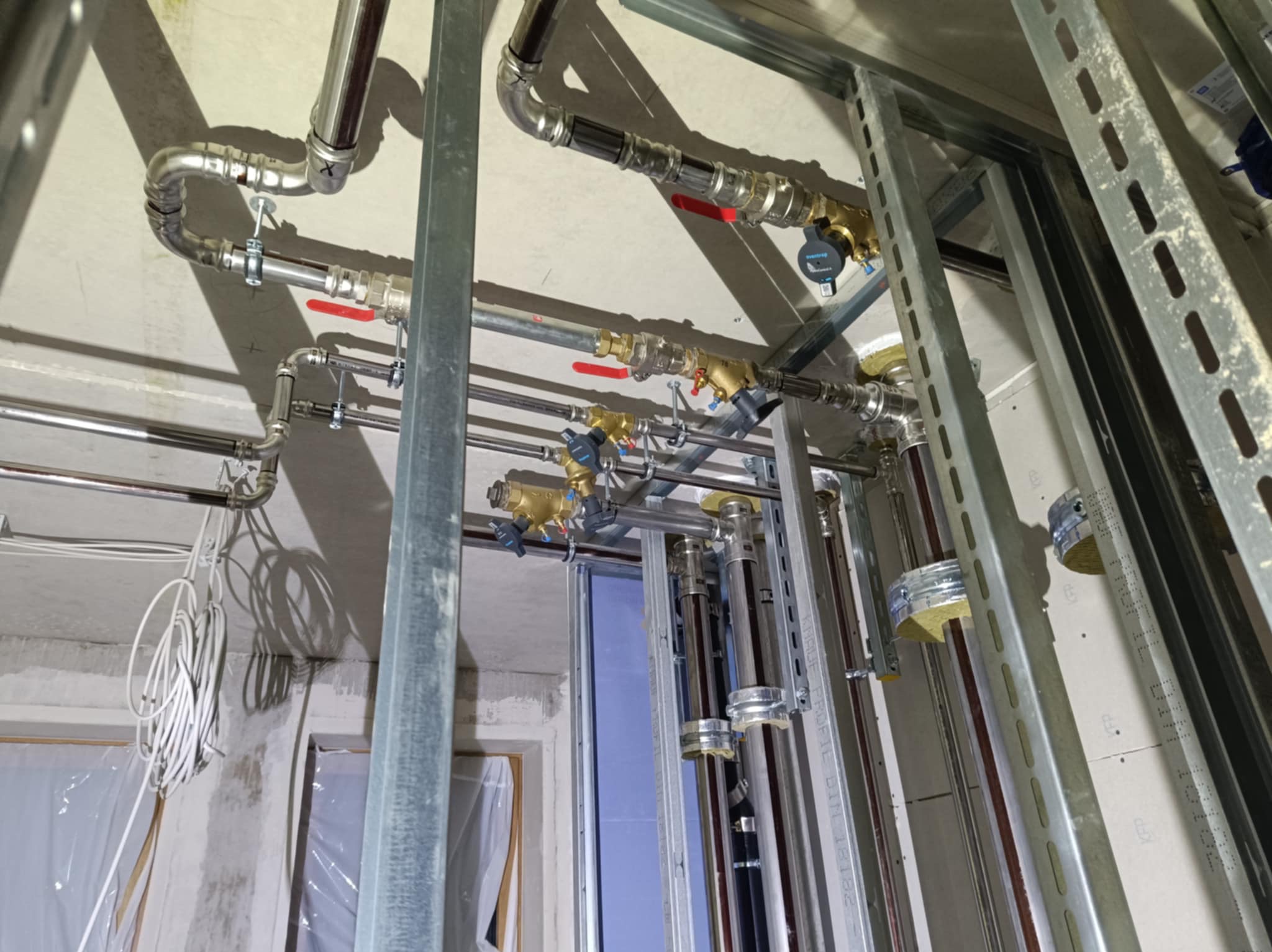 Widok z dołu na instalację hydrauliczną i grzewczą w trakcie montażu, z widocznymi rurami miedzianymi, zaworami z czerwonymi uchwytami, stalowymi profilami konstrukcyjnymi i zwisającymi kablami...