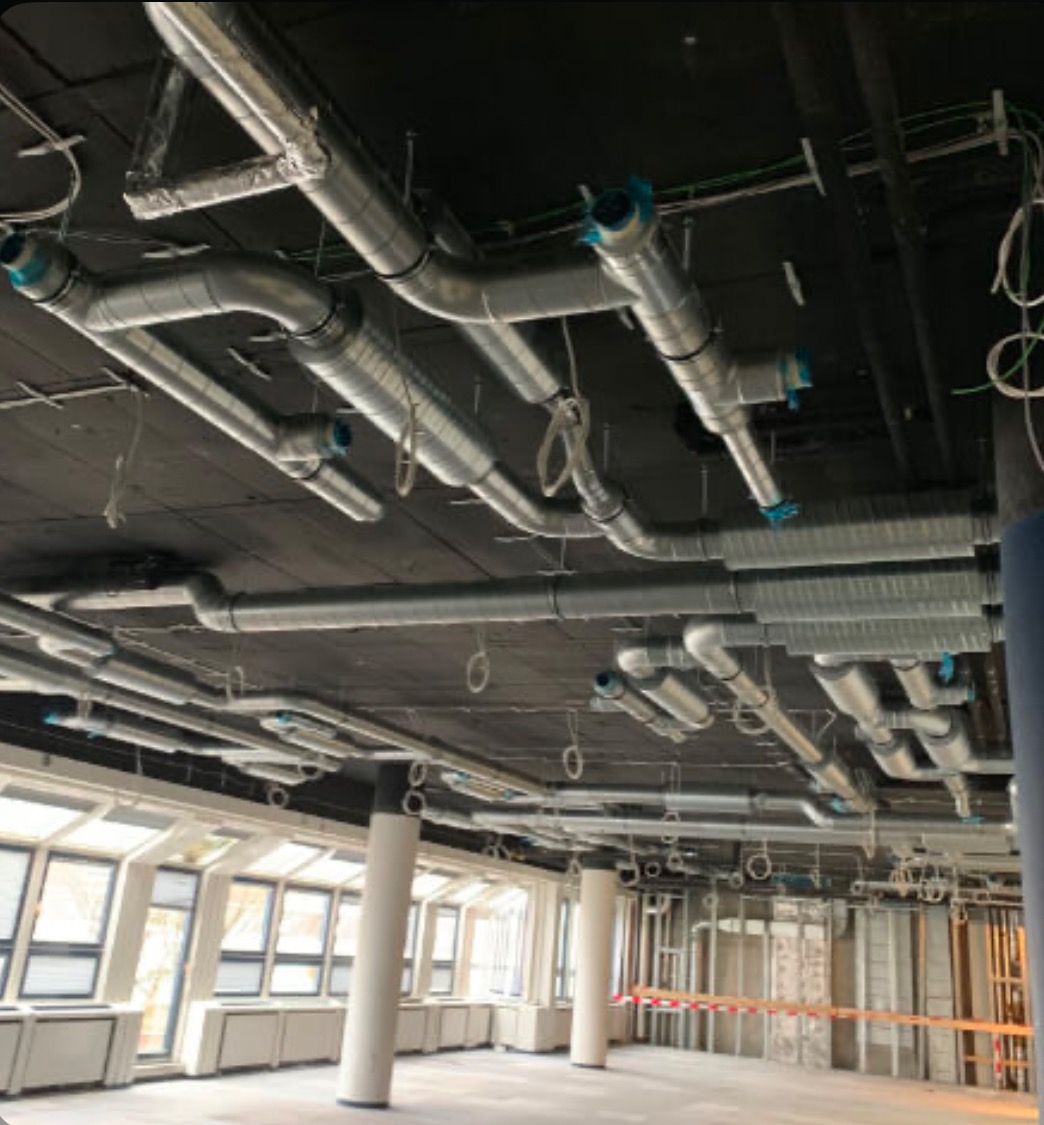 Skomplikowany system rur wentylacyjnych z niebieskimi elementami łączącymi, zamontowany pod czarnym sufitem w surowym wnętrzu biurowym z dużymi oknami i kolumnami.