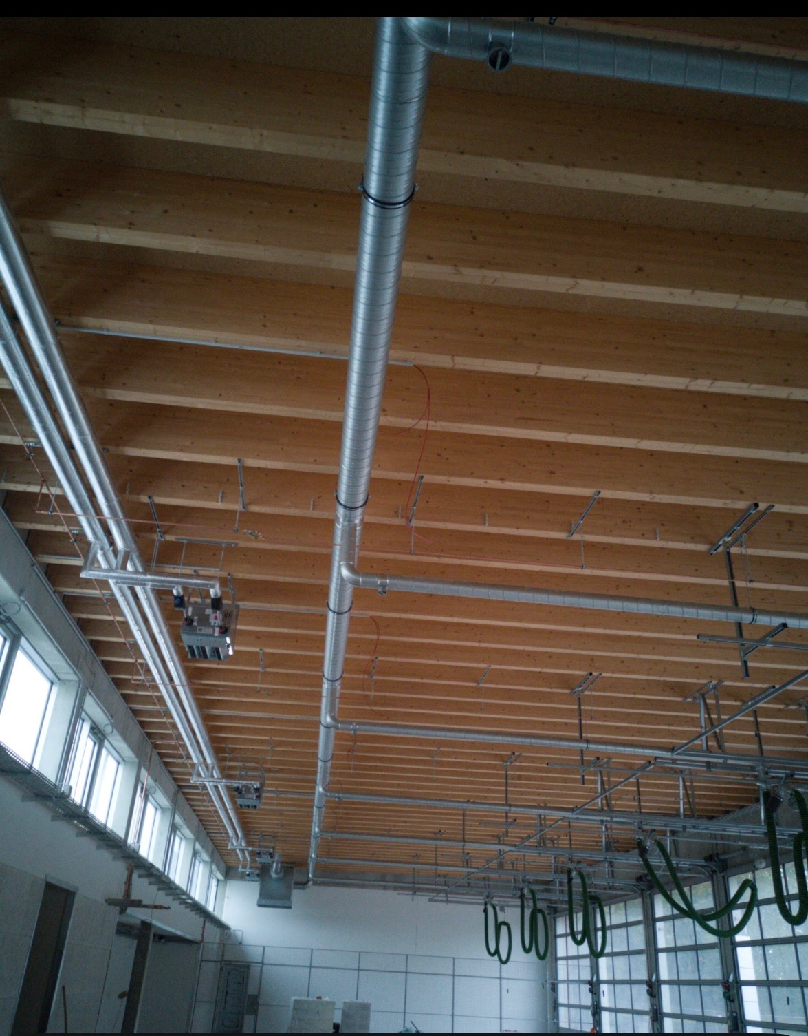 Metalowe rury wentylacyjne zamontowane pod drewnianym stropem w hali, widoczne okna i brama garażowa.