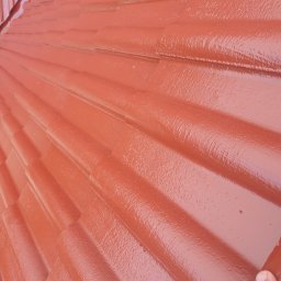 Arkadiusz Dorniak - Świeżo pomalowany dach z ceramicznej dachówki w odcieniach czerwieni, widoczna mokra, błyszcząca powierzchnia farby i fragment dłoni w prawym dolnym rogu.