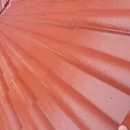 Świeżo pomalowany dach z ceramicznej dachówki w odcieniach czerwieni, widoczna mokra, błyszcząca powierzchnia farby i fragment dłoni w prawym dolnym rogu.