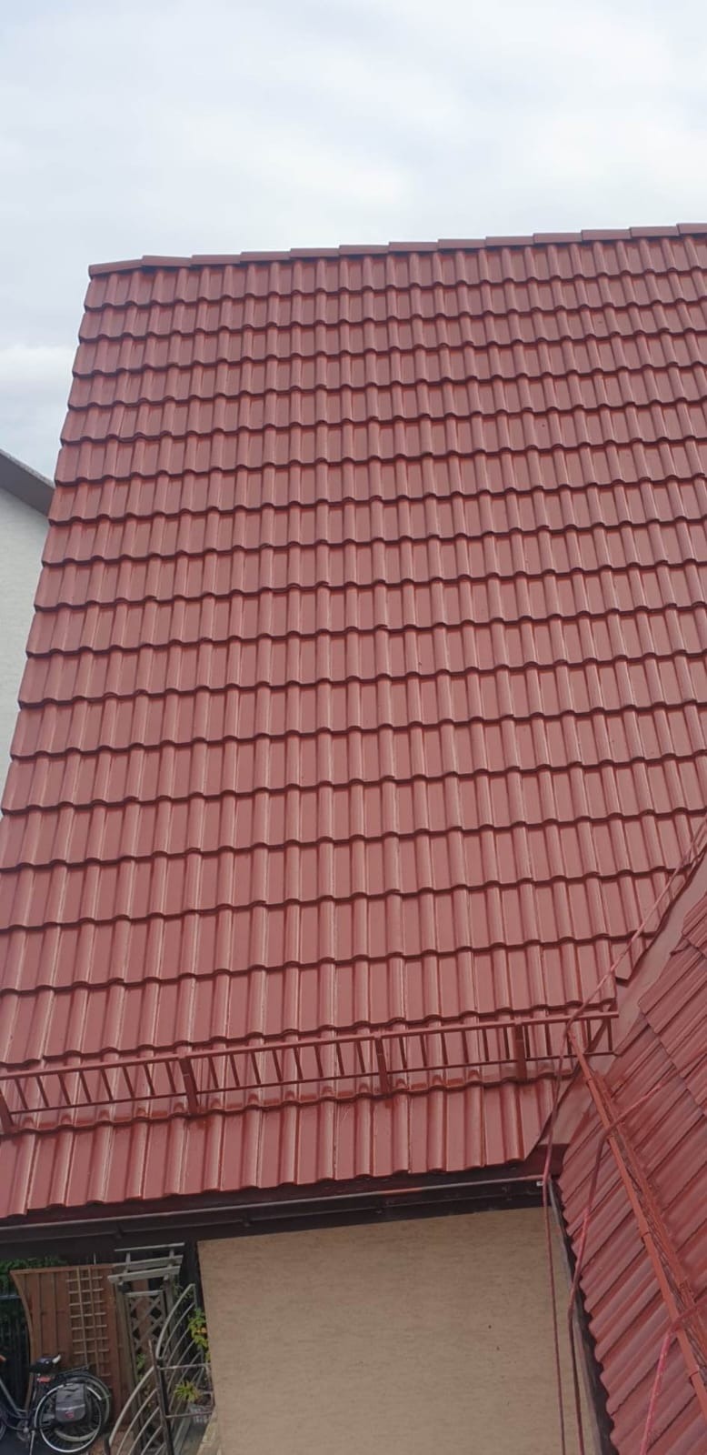 Czerwona dachówka ceramiczna na dachu domu jednorodzinnego, widok z dołu z fragmentem drabiny dekarskiej i rynny.