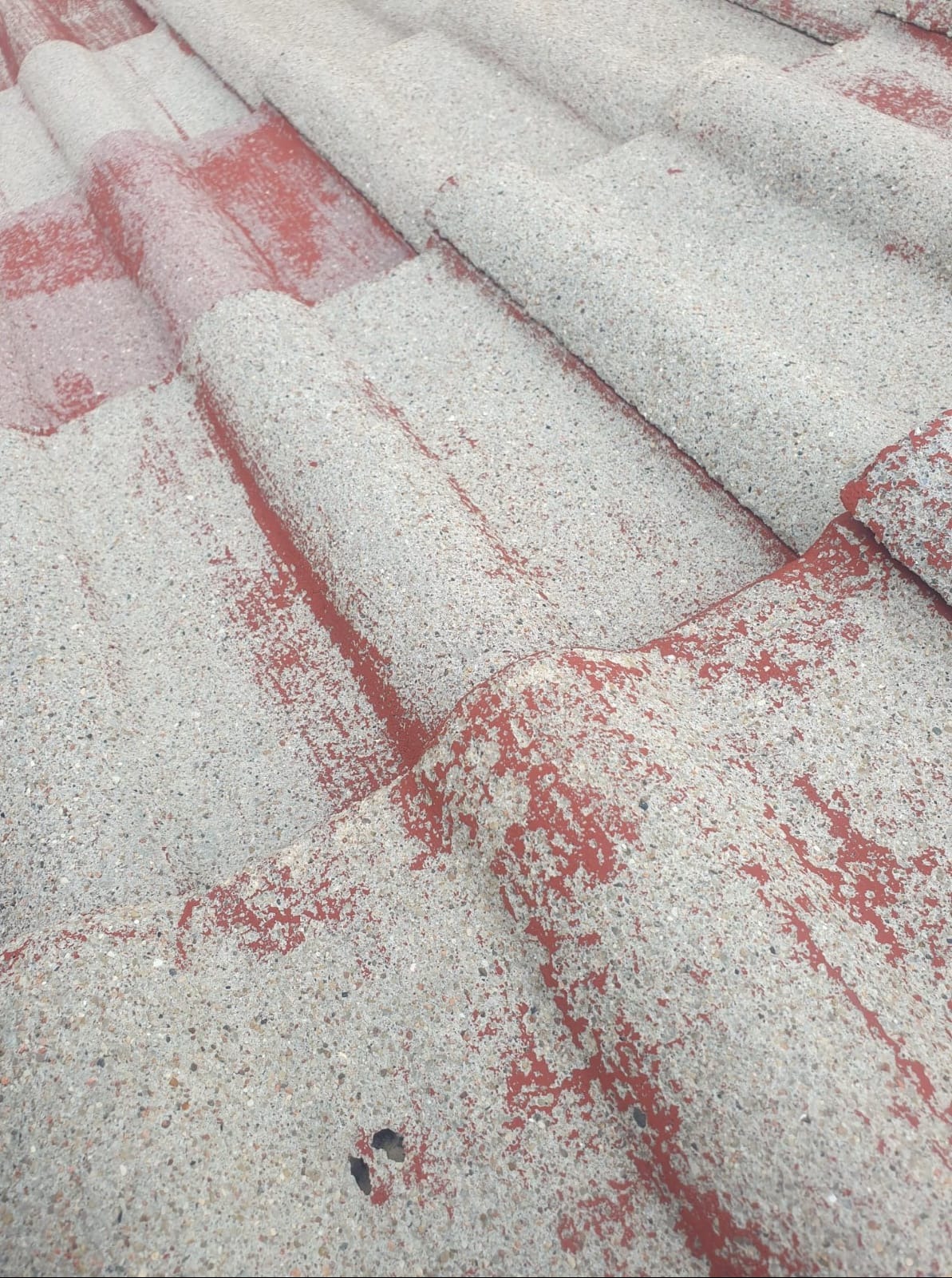 Niedokładnie pomalowana dachówka ceramiczna w kolorze czerwonym, z widocznymi szarymi fragmentami i porowatą strukturą.