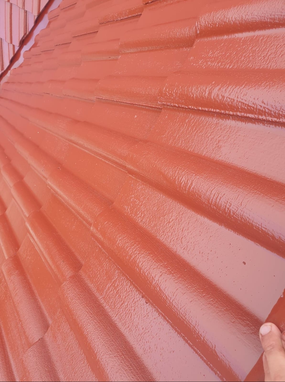 Świeżo pomalowany dach z ceramicznej dachówki w odcieniach czerwieni, widoczna mokra, błyszcząca powierzchnia farby i fragment dłoni w prawym dolnym rogu.