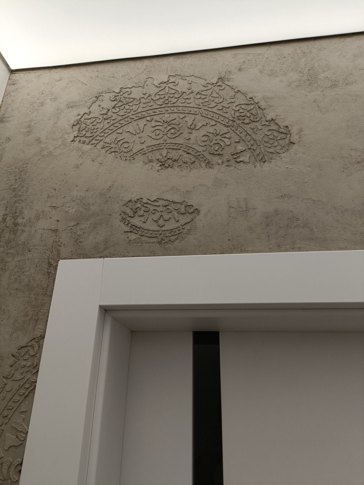 Dekoracyjna ściana z szarego tynku strukturalnego z ornamentami w stylu arabskim, widoczny fragment białej futryny drzwiowej z czarnym elementem dekoracyjnym.