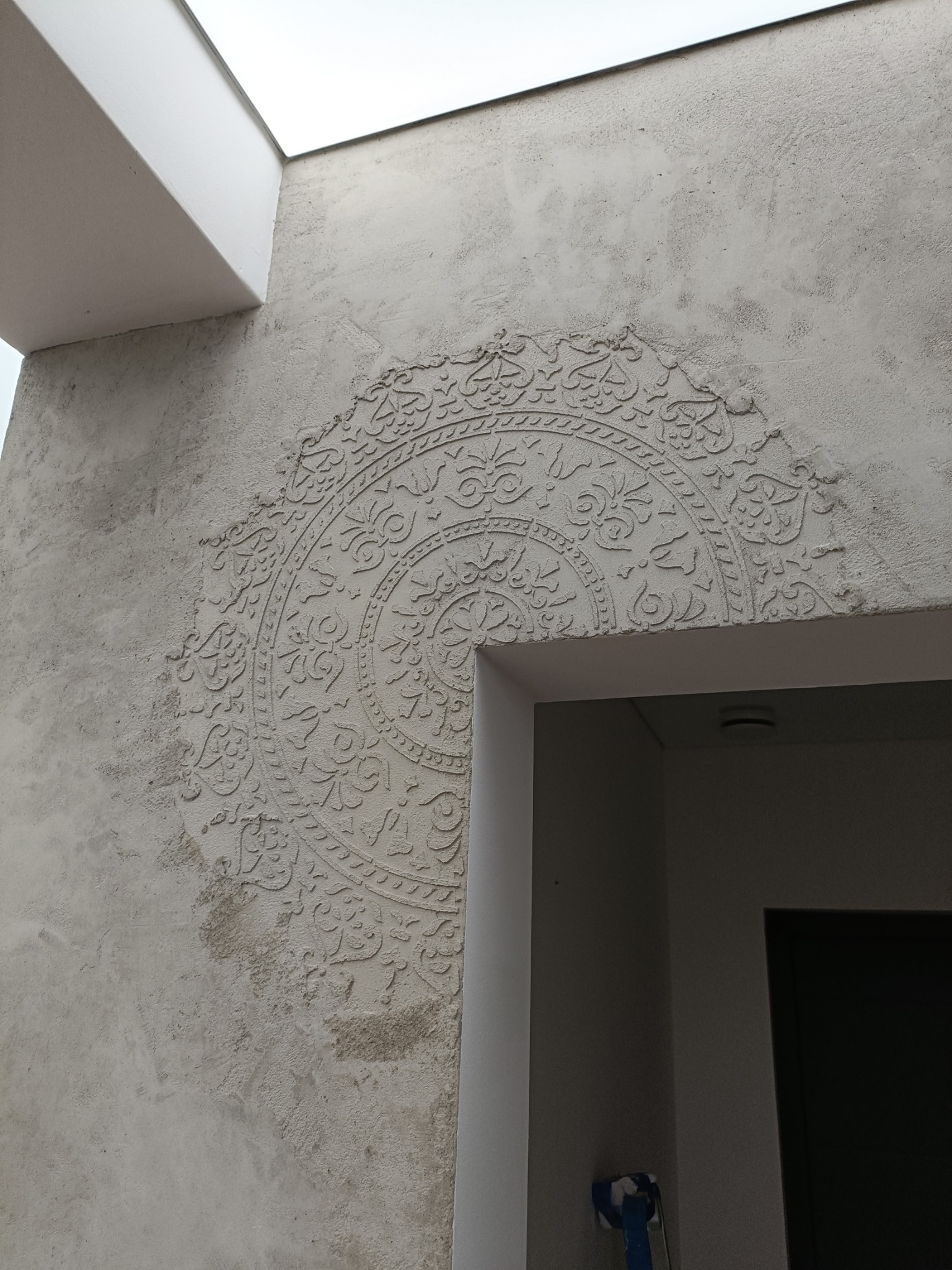 Dekoracyjna ściana z ornamentem w kształcie mandali, wykonana techniką reliefu na szarej, teksturowanej powierzchni, widoczna obok białego obramowania drzwi i fragmentu sufitu.