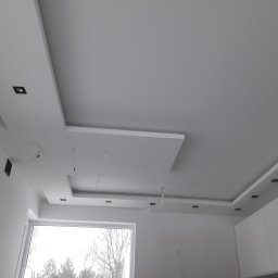 Usługi Remontowo-Budowlane Adam Ciak - Biały sufit podwieszany z wbudowanymi czarnymi punktami świetlnymi, widoczne kable zwisające z sufitu, okno z widokiem na drzewa bez liści, minimalistyczny design wnętrza.