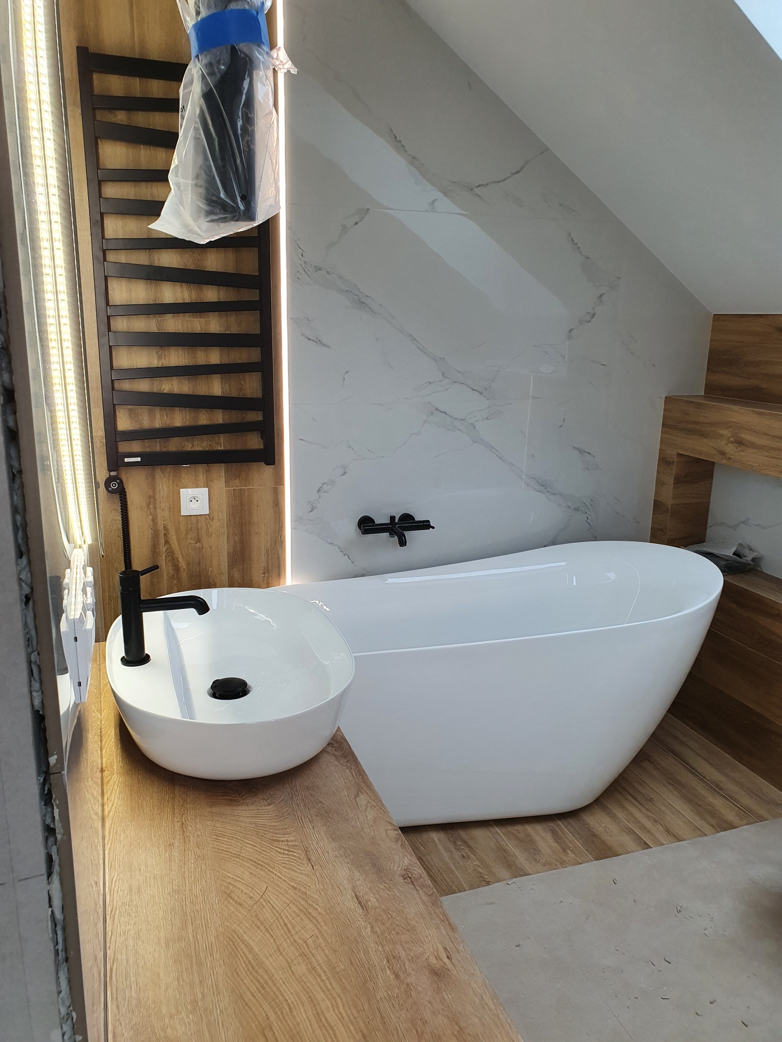 Nowoczesna łazienka na poddaszu z wolnostojącą wanną, umywalką nablatową i grzejnikiem drabinkowym w folii ochronnej, marmurowe płytki na ścianie.