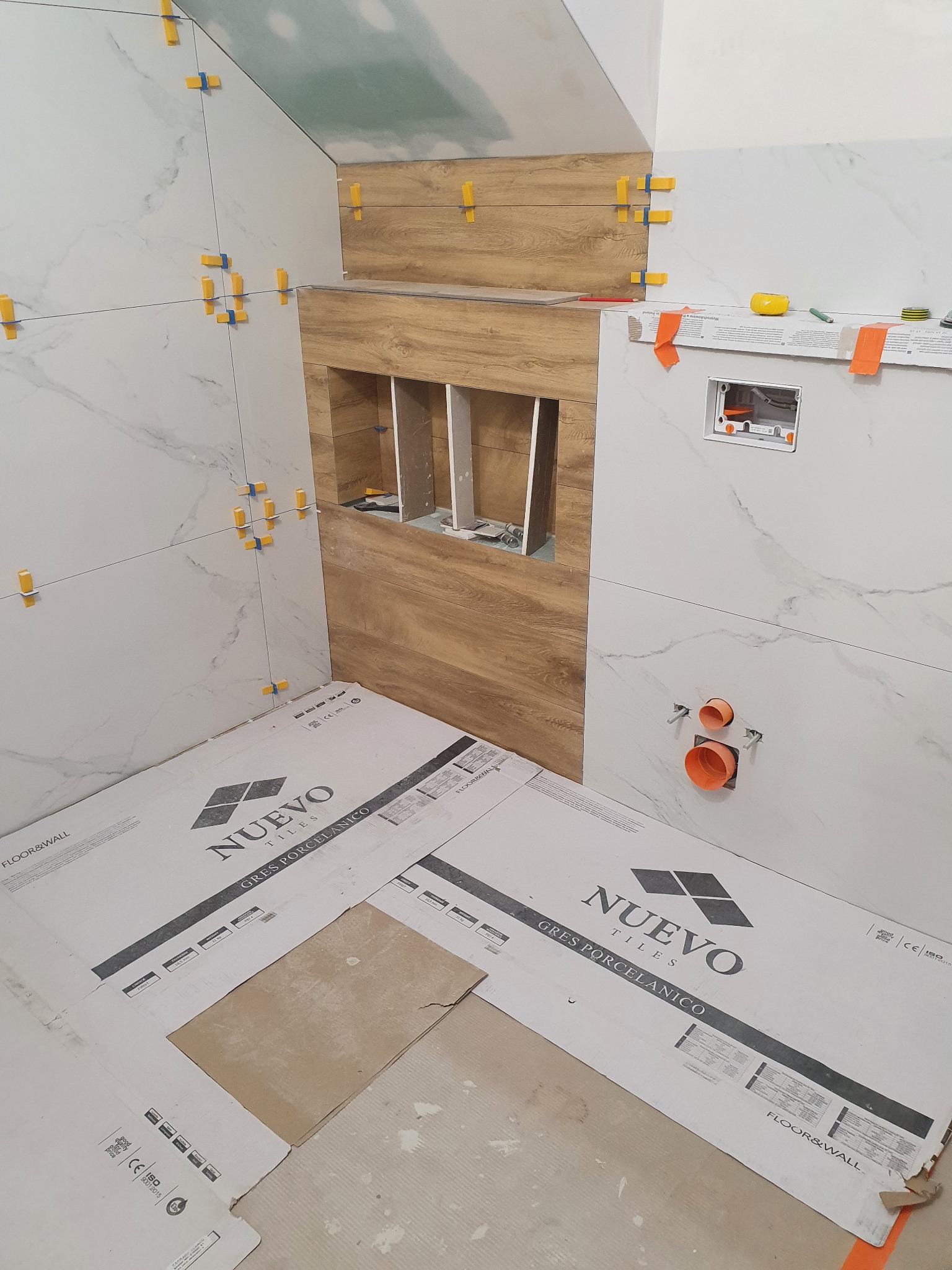 Montaż płytek imitujących drewno w łazience w Olszewnicy Starej, widoczne listwy dystansowe, wnęka z półkami w trakcie wykańczania, zabezpieczone podłoże kartonami z logo producenta płytek.