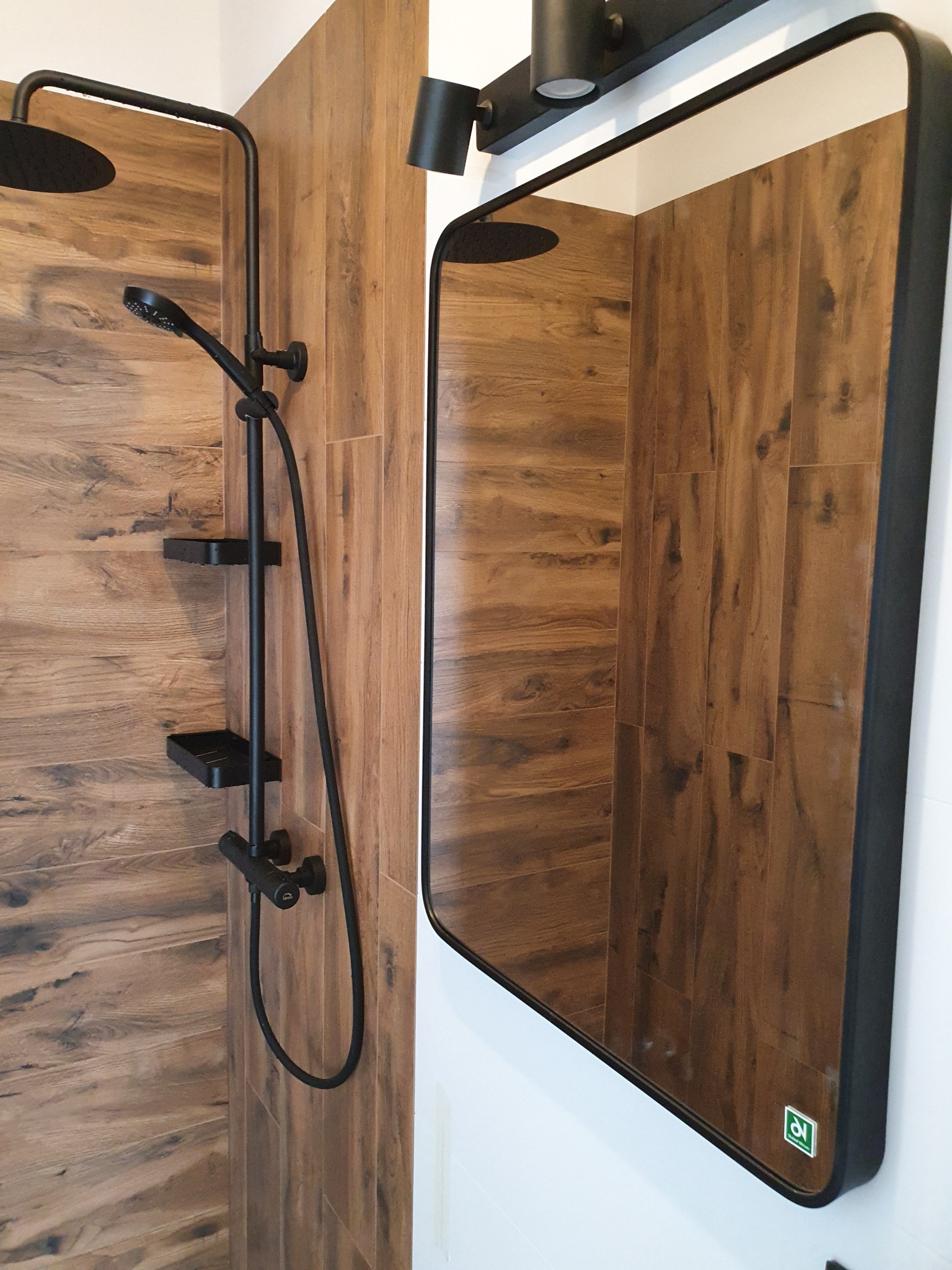 Nowoczesna łazienka z kabiną prysznicową typu walk-in, wykończona płytkami imitującymi drewno, z czarną armaturą i prostokątnym lustrem w czarnej ramie.