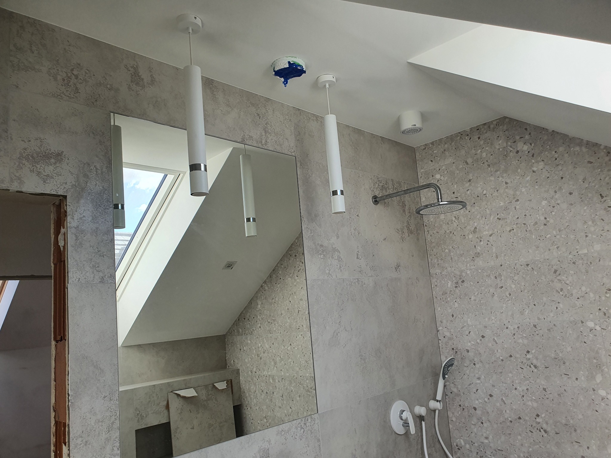 Wnętrze łazienki w trakcie wykańczania, widoczne płytki imitujące lastryko na ścianach, nowoczesna armatura prysznicowa, białe lampy wiszące, lustro odbijające okno dachowe oraz zabezpieczone przed...