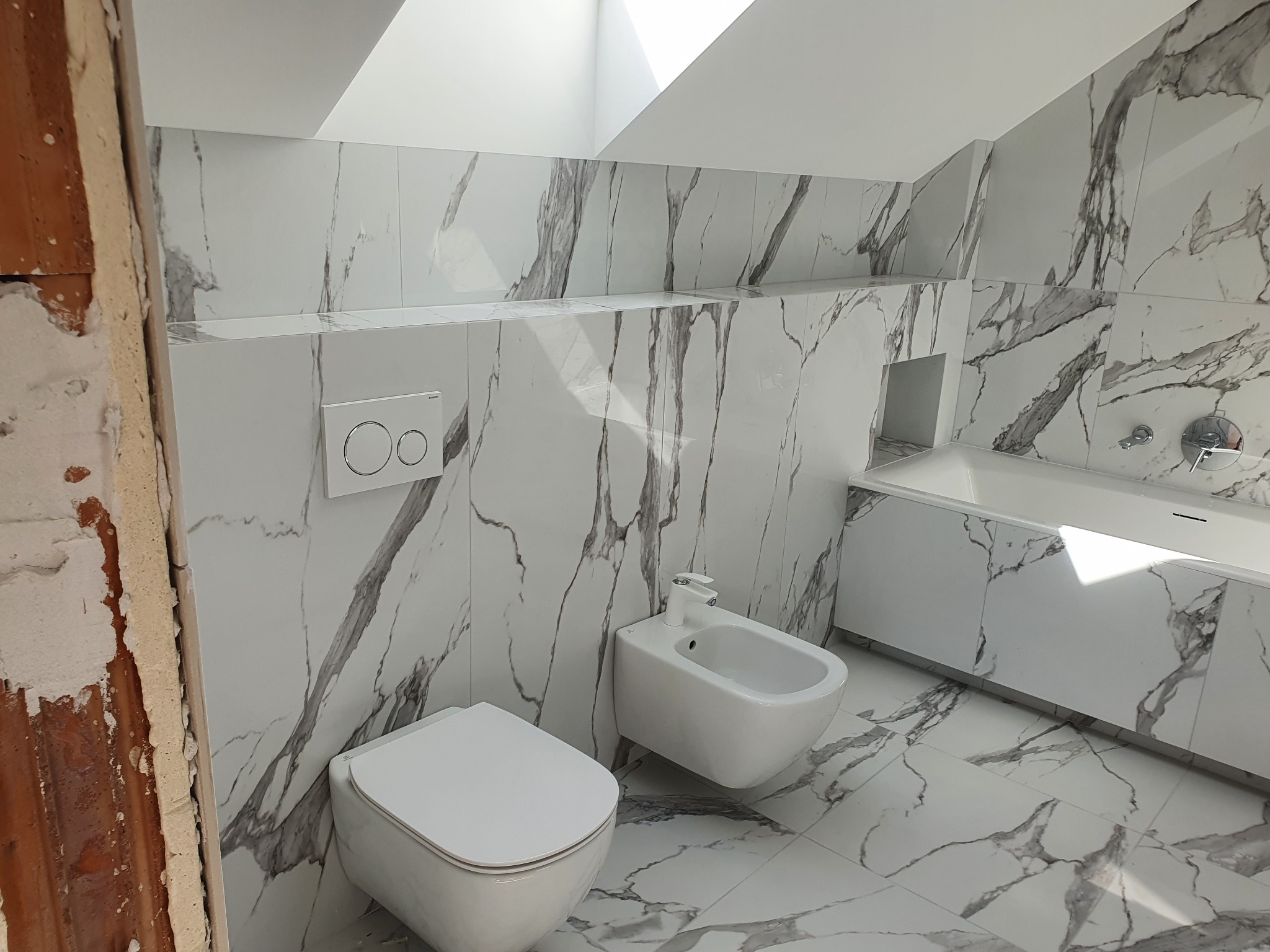 Nowoczesna łazienka w bieli i szarościach z marmurowym wzorem na płytkach, widok na toaletę, bidet i zabudowaną wannę, okno dachowe wpuszcza naturalne światło, surowa ściana z cegły po lewej stronie.