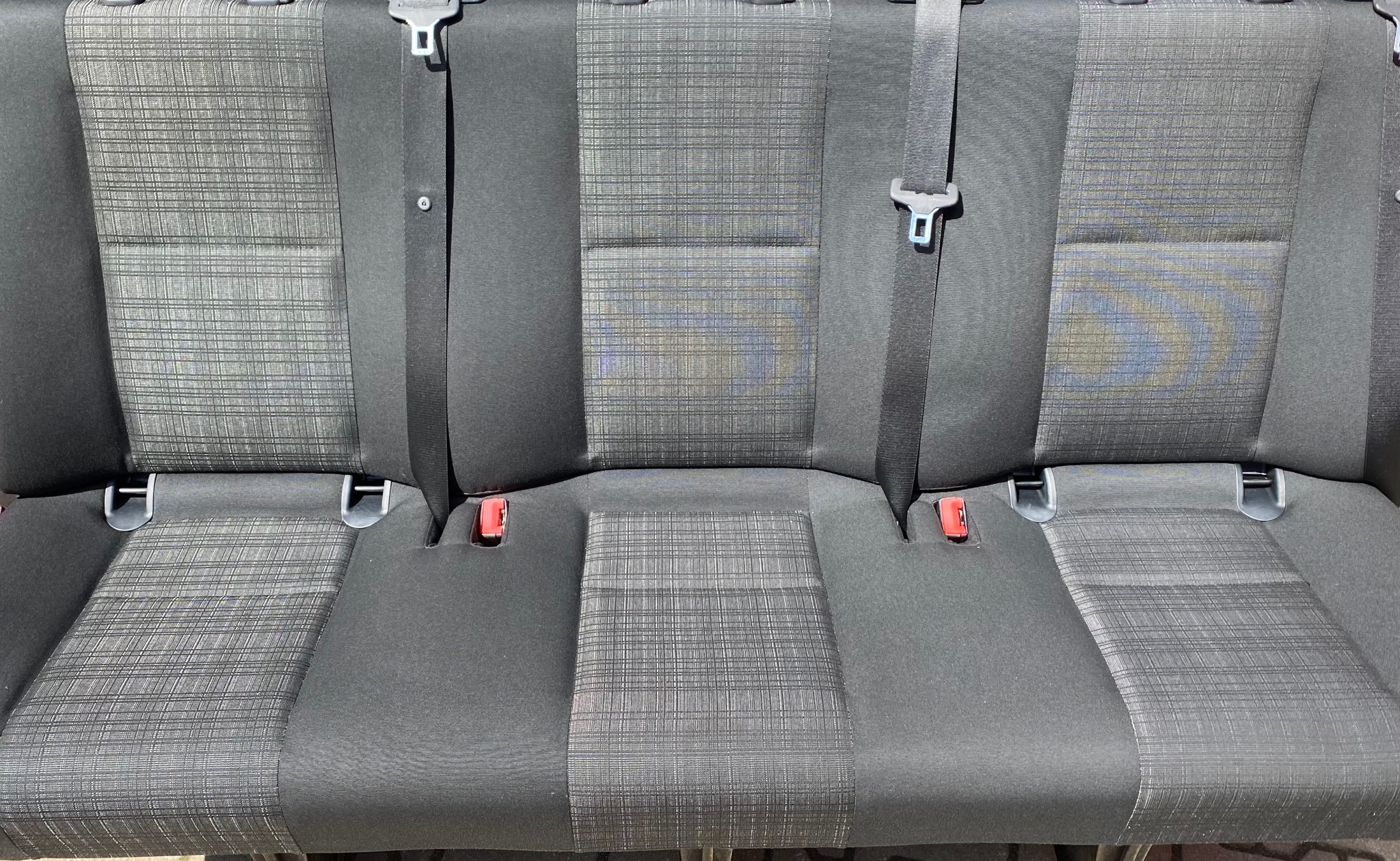 Tapicerka samochodowa z plamą na środkowym siedzeniu, widoczne pasy bezpieczeństwa i mocowania. Tekstylna faktura materiału.