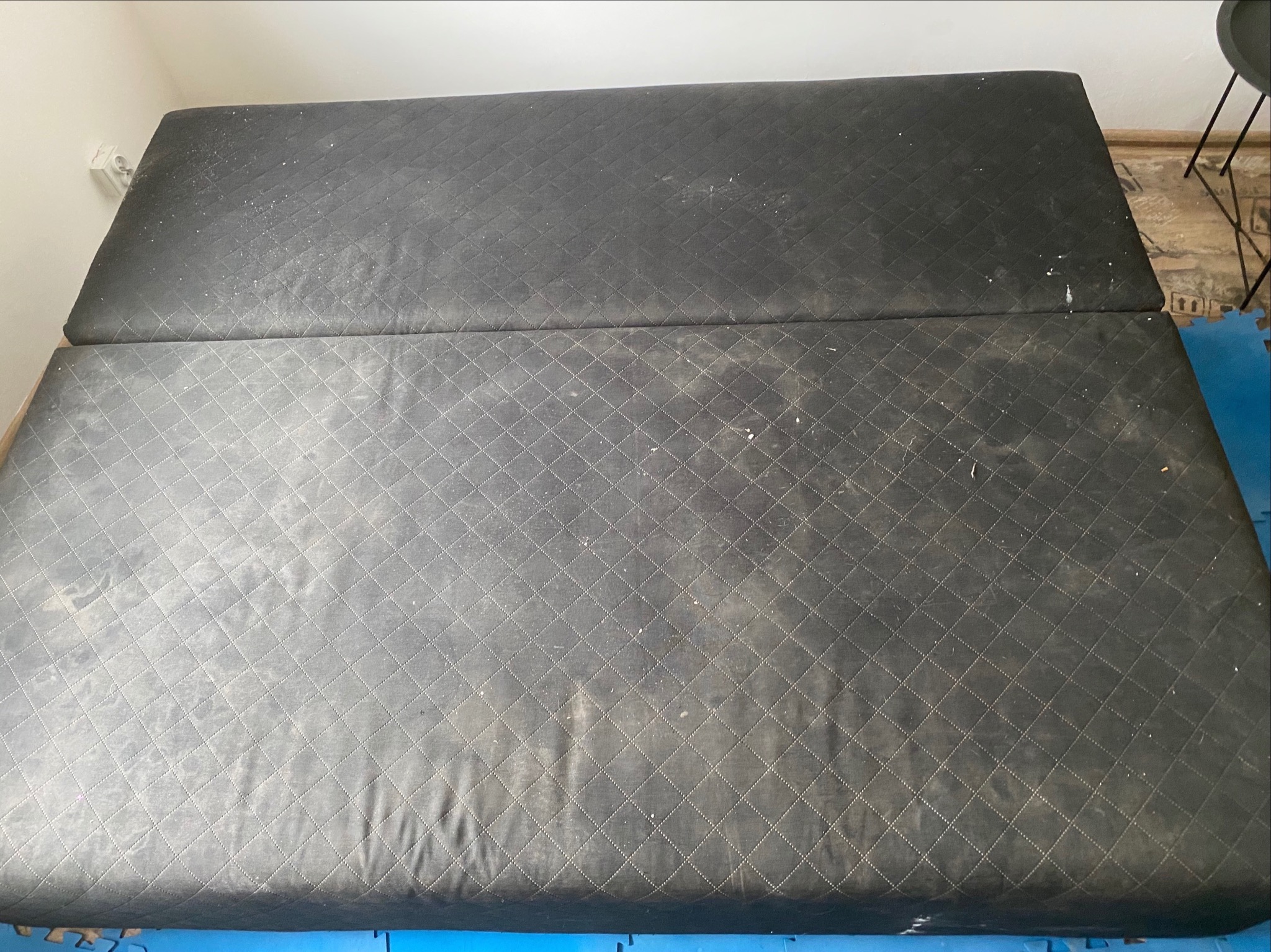 Ciemnoszary materac z pikowaniem w romby, widoczne zabrudzenia i plamy, sugerujące potrzebę czyszczenia tapicerki. Ujęcie z góry, widoczny fragment podłogi.