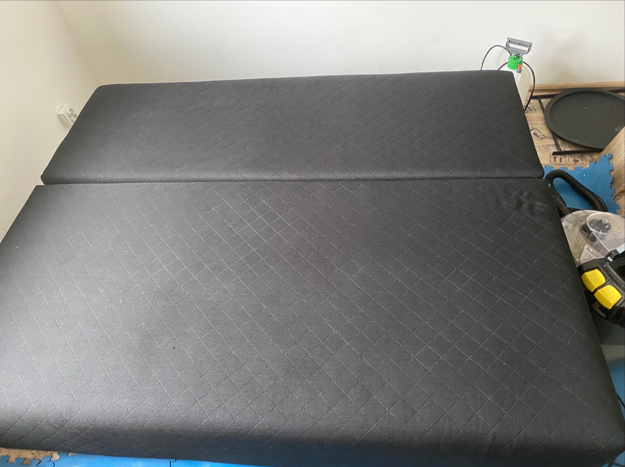 Ciemnoszary, pikowany materac z widocznymi narzędziami do czyszczenia, sugerujący etap po praniu tapicerki. Widok z góry, bez osób.